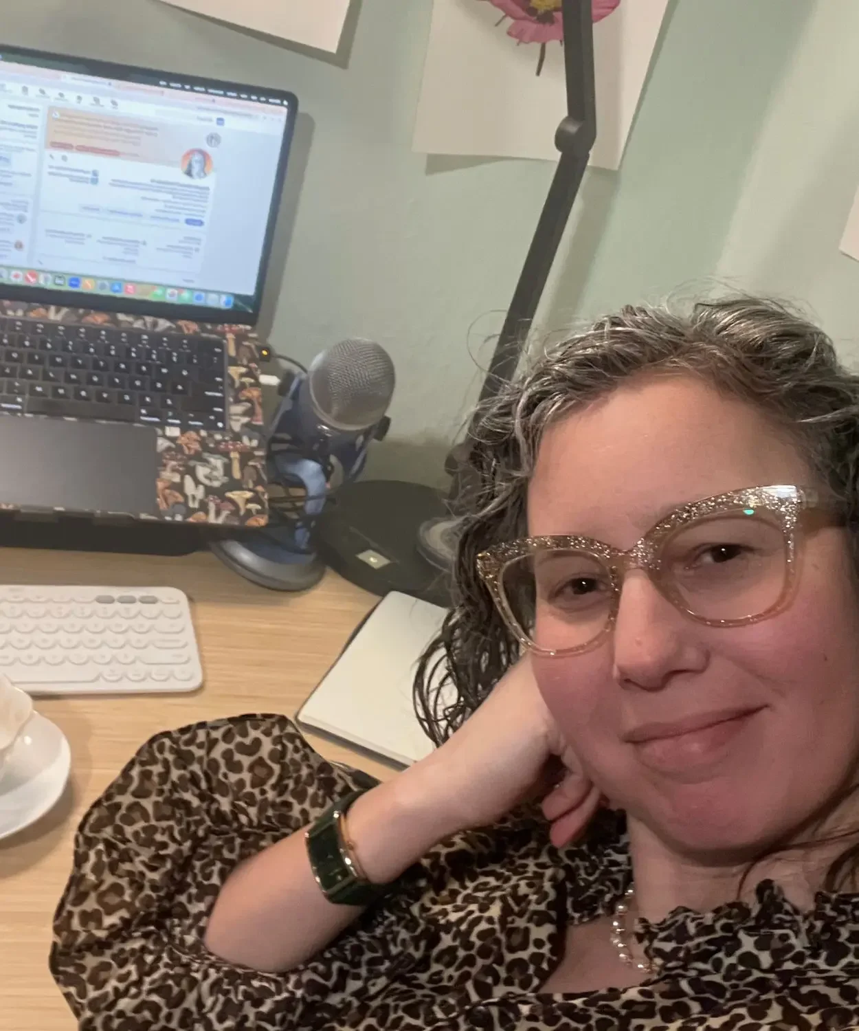 Angela Marie D’Antonio, education keynote speaker, sitting at her desk with a laptop and microphone, wearing gold glitter glasses and a patterned blouse, smiling toward the camera in a home office workspace.