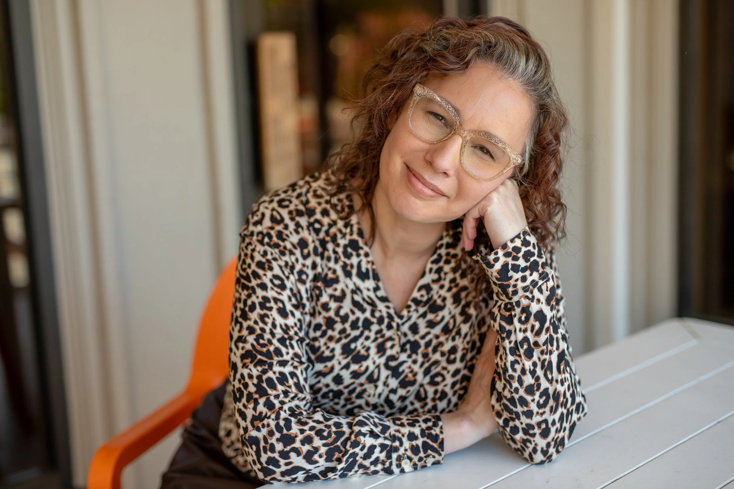 Angela Marie D'Antonio, keynote speaker and executive coach.  Image shows a woman with curly hair wearing big, sparkly glasses and a leopard-print blouse, smiling and resting her head on her hand while sitting at a white table.