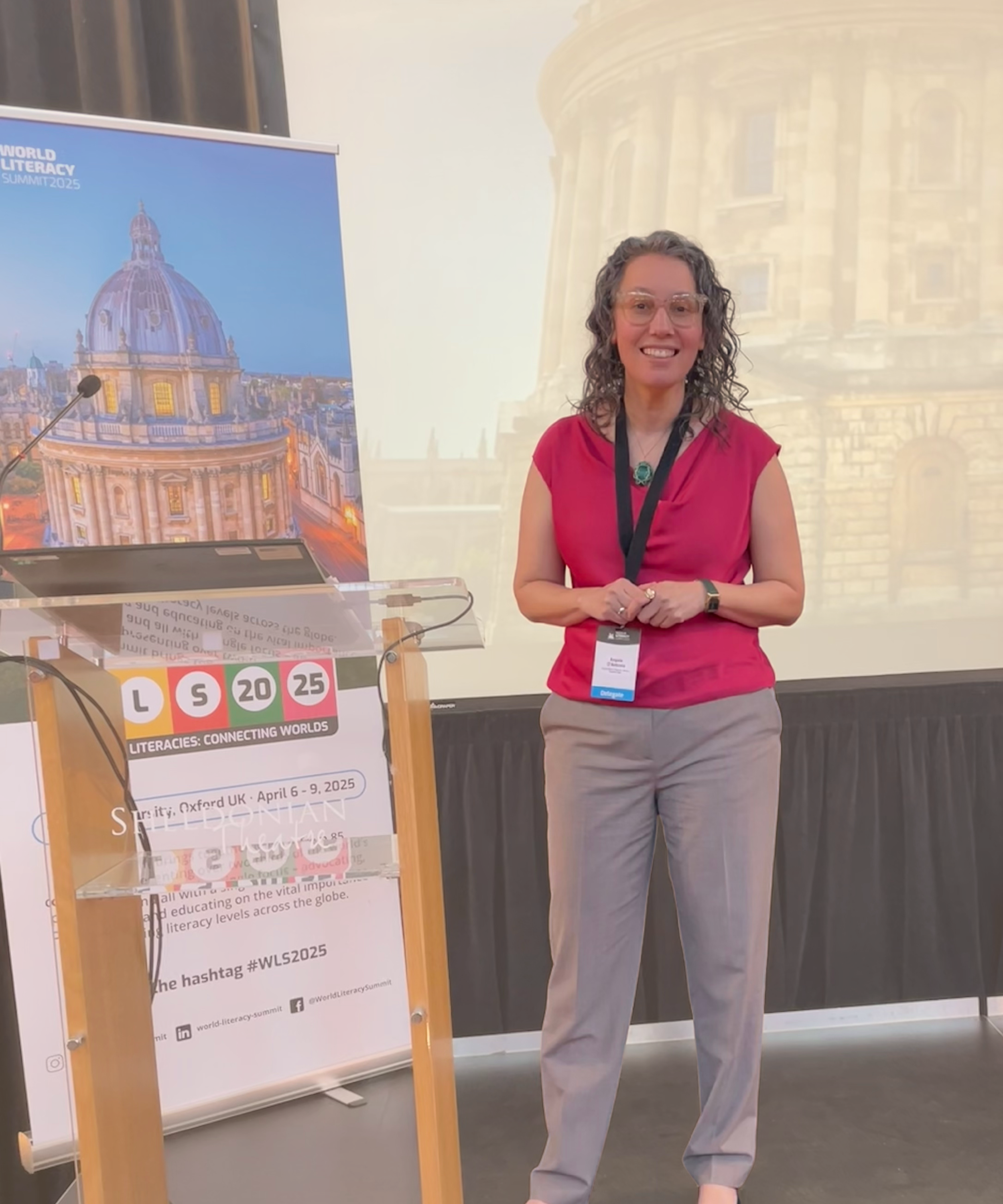 Angela Marie D'Antonio standing on a stage at the World Literacy Summit 2025, smiling, wearing a pink top, beige pants, and glasses, with a conference badge, behind a lectern with a poster featuring an architectural photo of Oxford University.