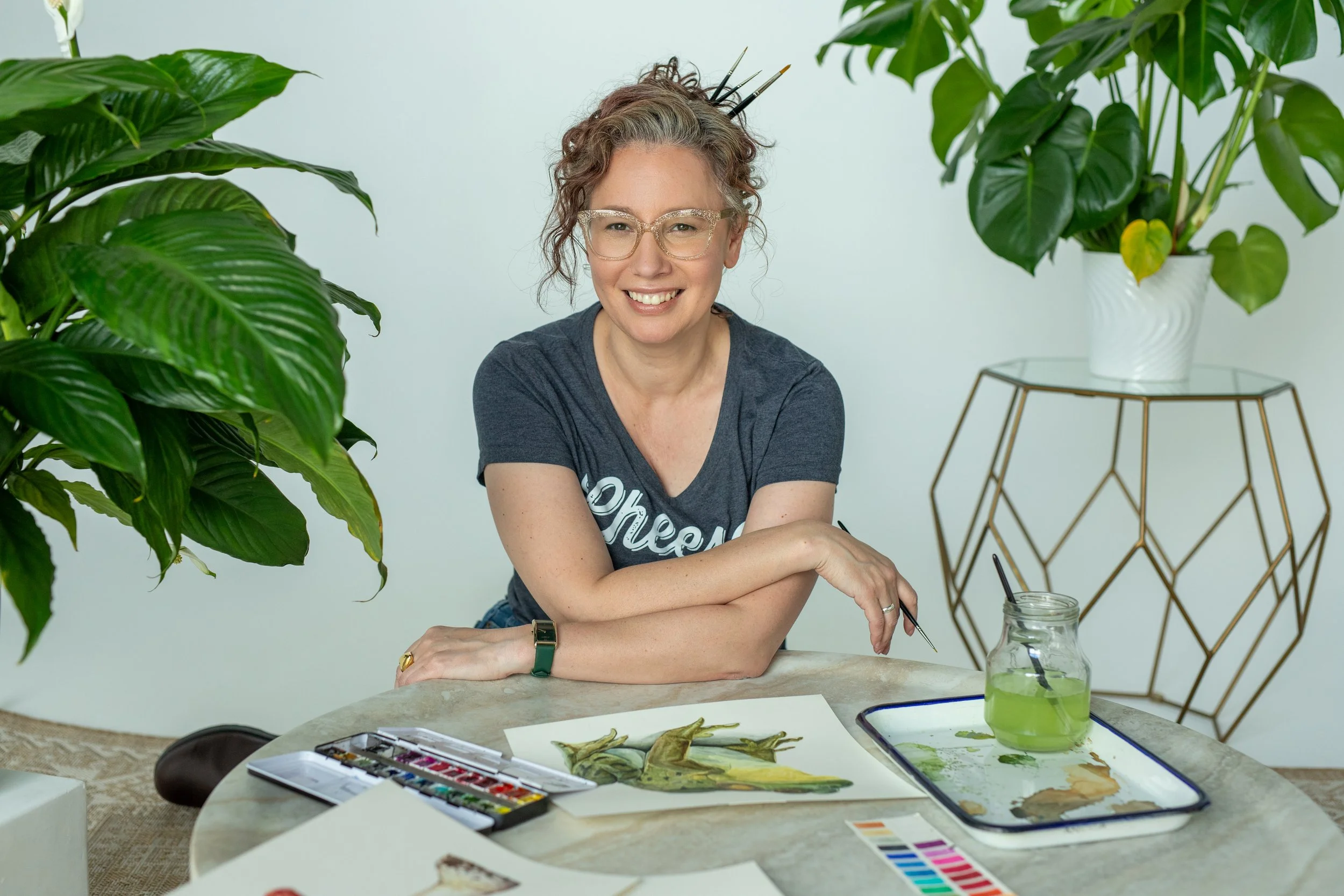 Angela Marie D'Antonio, executive coach for neurodivergent founders and weekend watercolorist, smiling at her painting table with brushes, paints, and her hyperrealistic nature paintings.