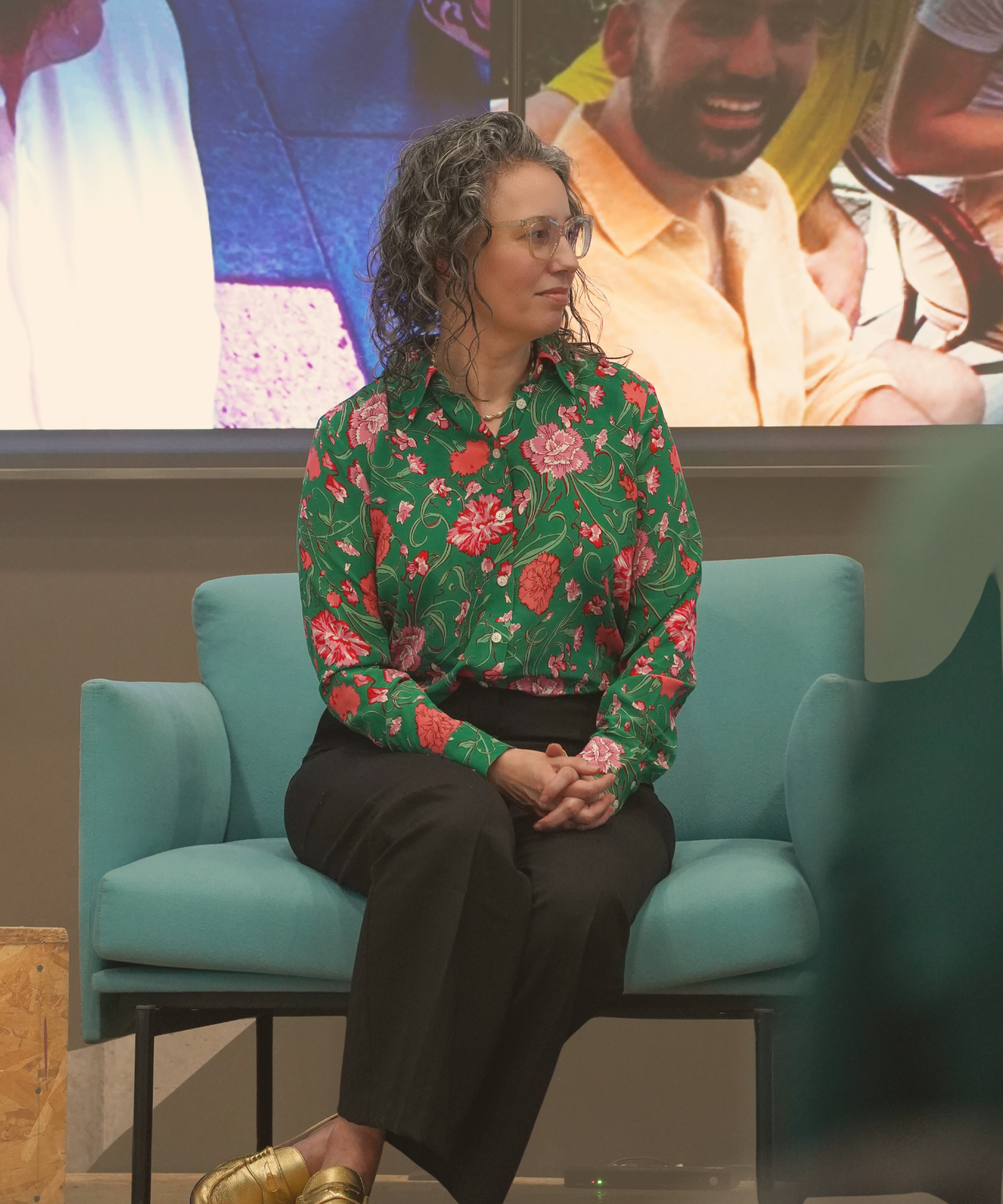 Angela Marie D'Antonio, a woman with curly hair and glasses sitting on a light blue chair, wearing a green floral blouse and black pants, in front of a large screen displaying images of people on a speaking panel at the LinkUp MeetUp event in Bosnia.
