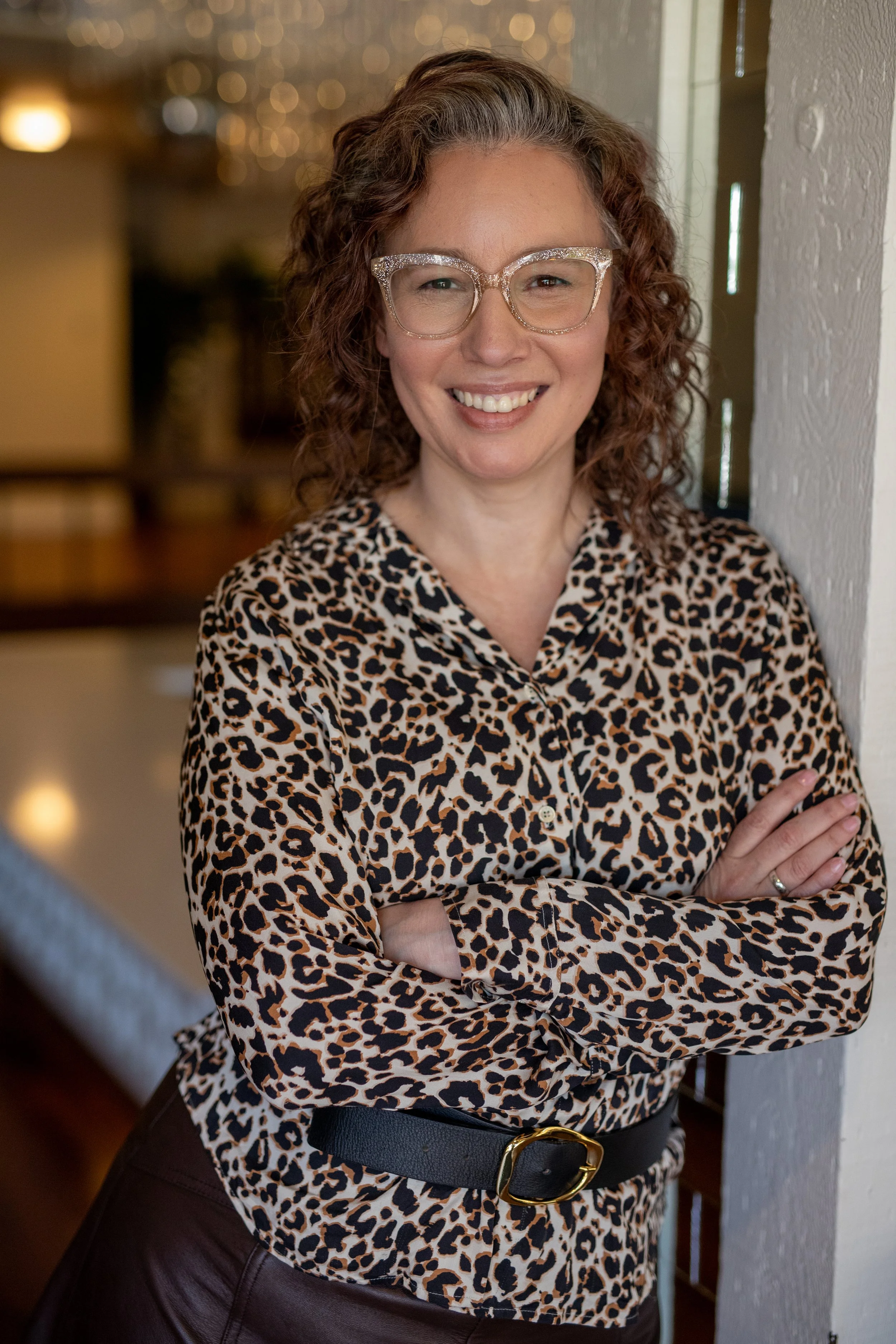Angela Marie D'Antonio, executive coach for neurodivergent founders and keynote speaker, smiling in clear glitter glasses and a leopard-print blouse with arms crossed in a warmly lit lobby.