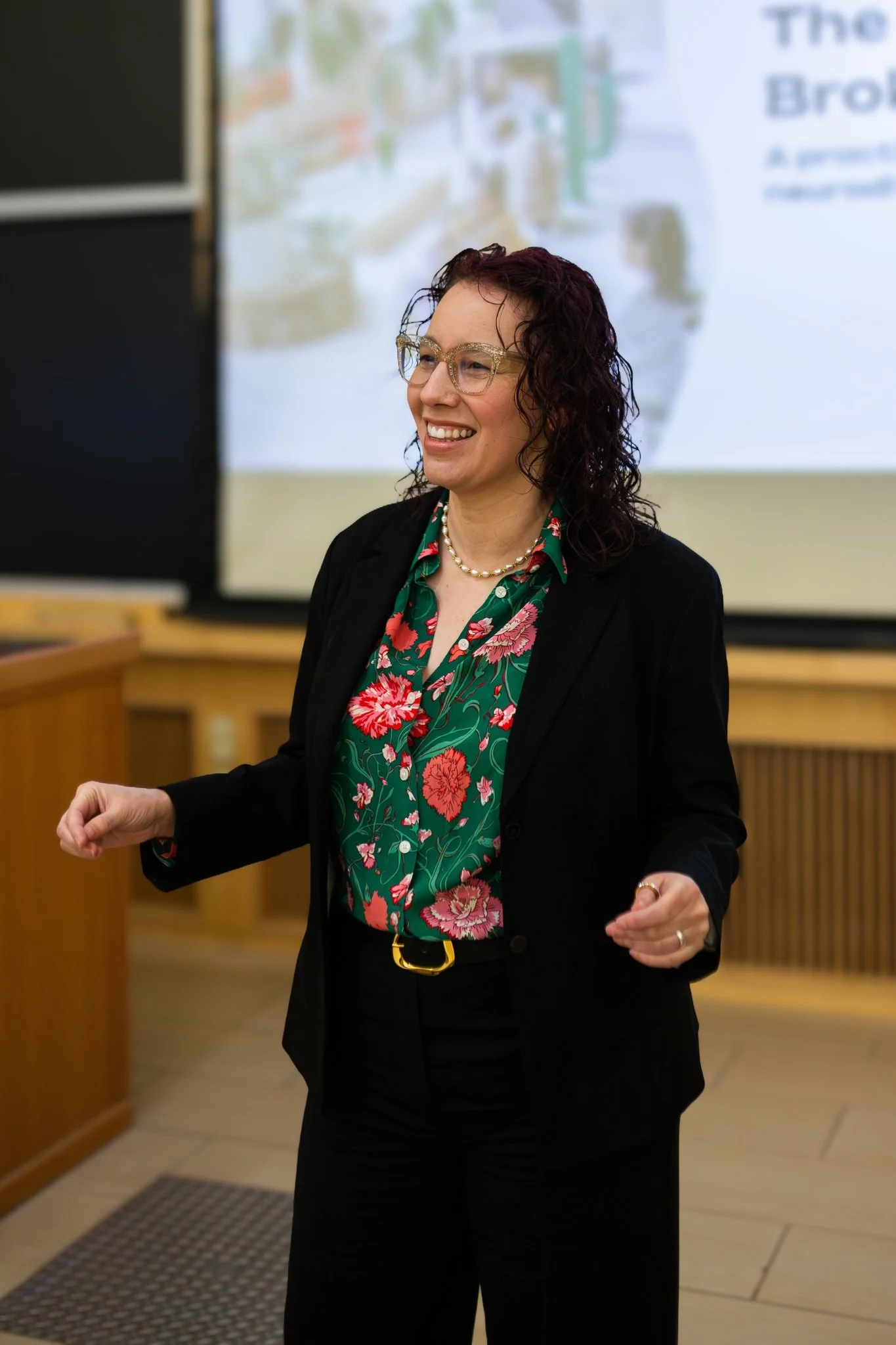 Angela Marie D'Antonio, keynote speaker and executive coach, presenting on supporting neurodivergent employees.  Image shows a woman with curly dark hair, gold glitter glasses, a floral green blouse, and a black blazer speaking in a conference room.