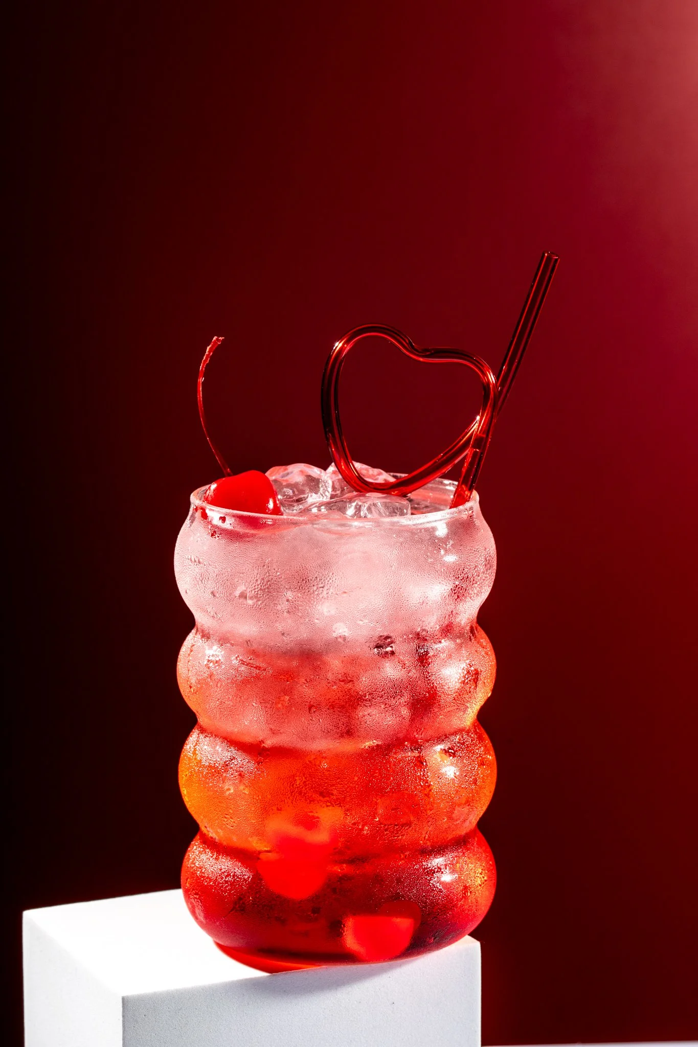 dirty shirley temple sits on the edge of a white block with cherry juice and maraschino balanced on top on a red background