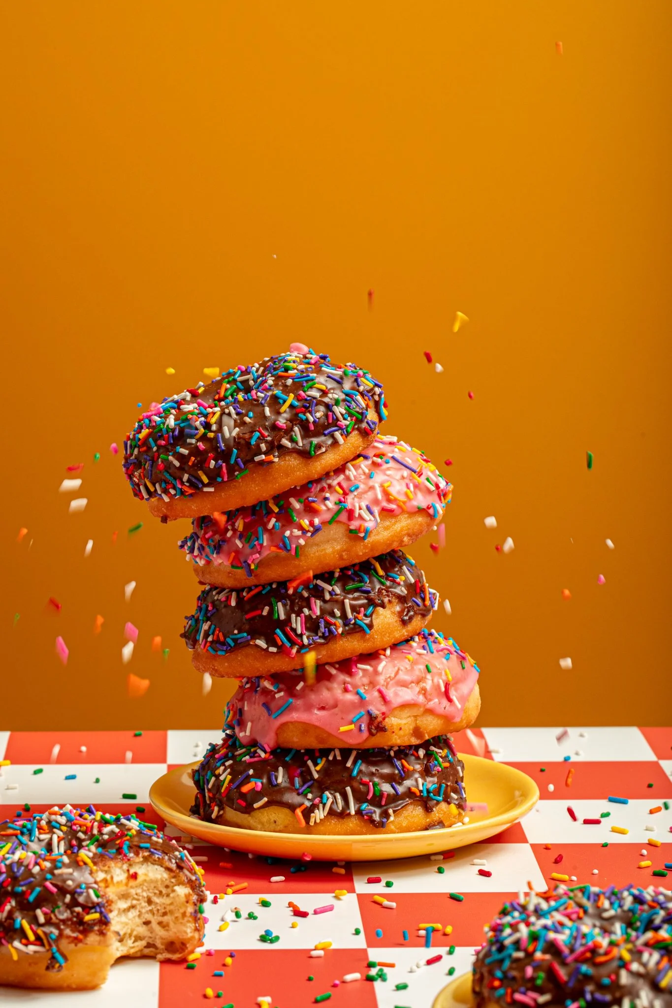 pile of colorful donuts stacked high about to fall over as sprinkles rain down on top donut photos on yellow background