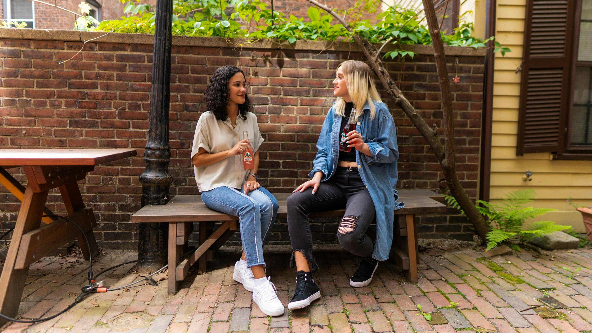 Two women sitting on a wooden bench in a backyard, smiling and talking while holding bottles of soda. The backyard has a brick wall, some greenery, and a yellow house with open shutters.