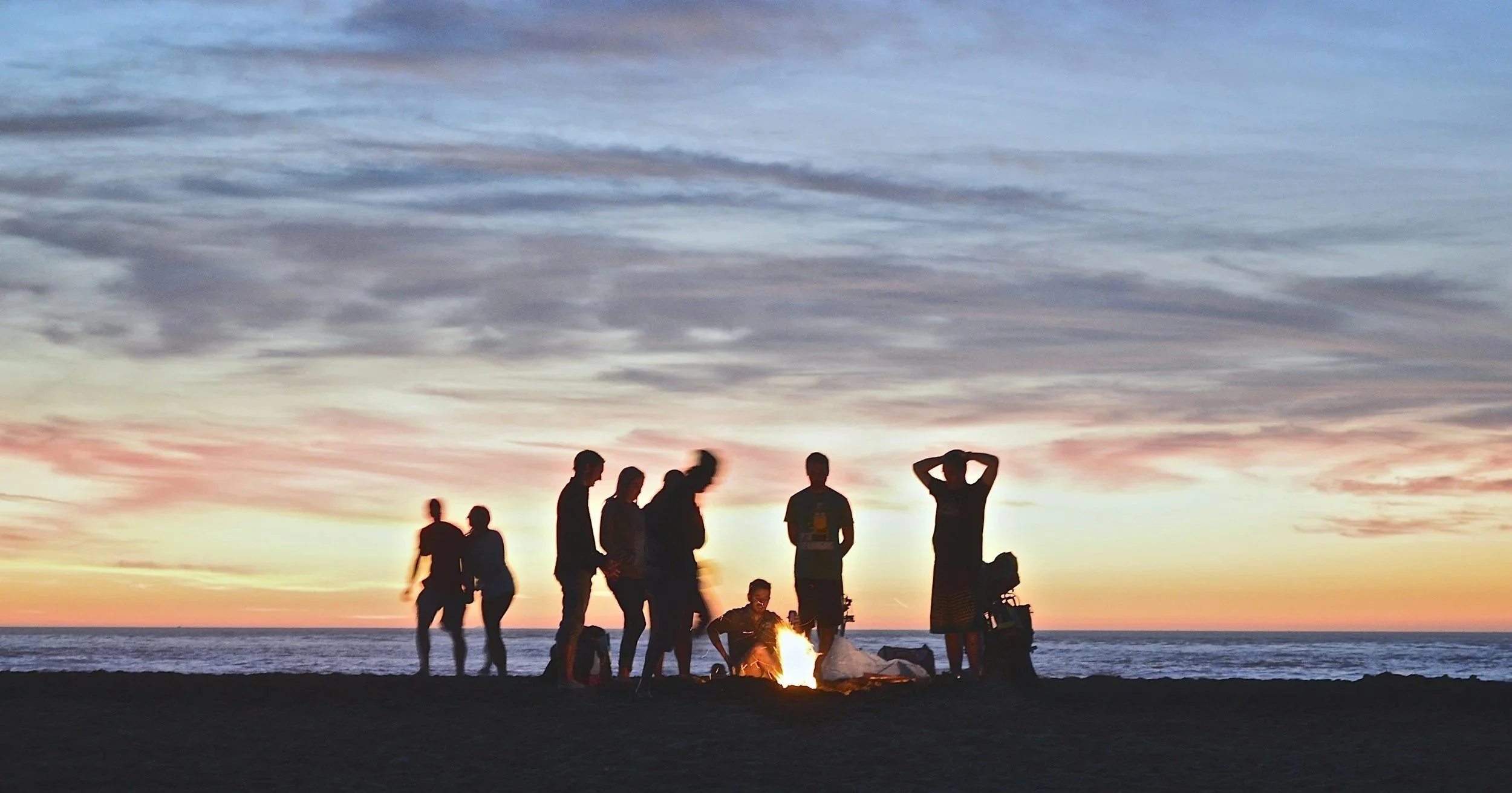 Group of friends gathered around a bonfire on the beach at sunset, representing community