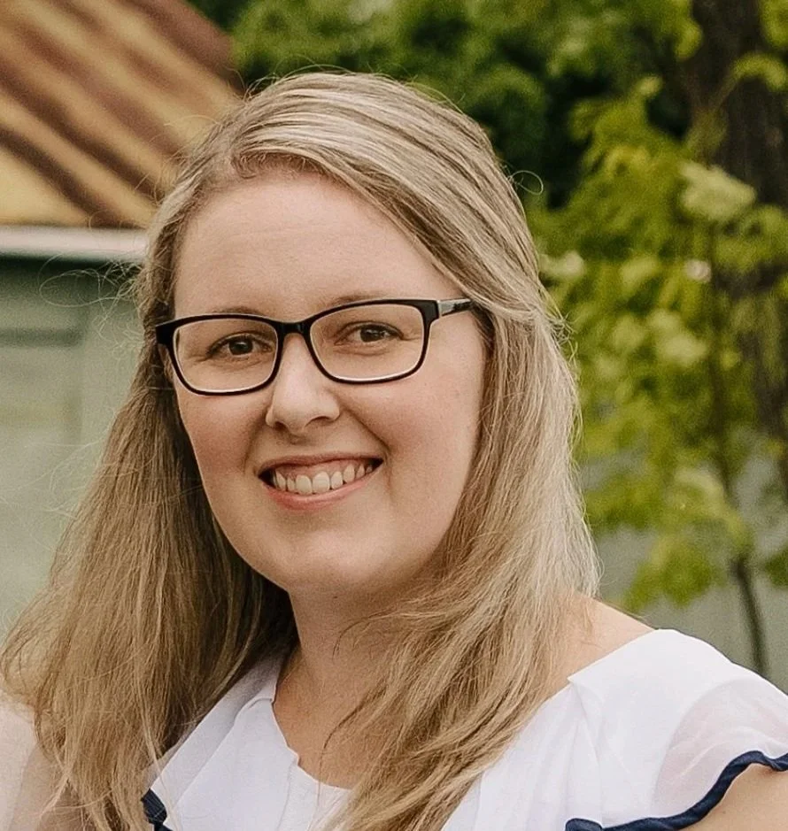 A smiling woman with long blonde hair and glasses outdoors, with a house and green trees in the background.