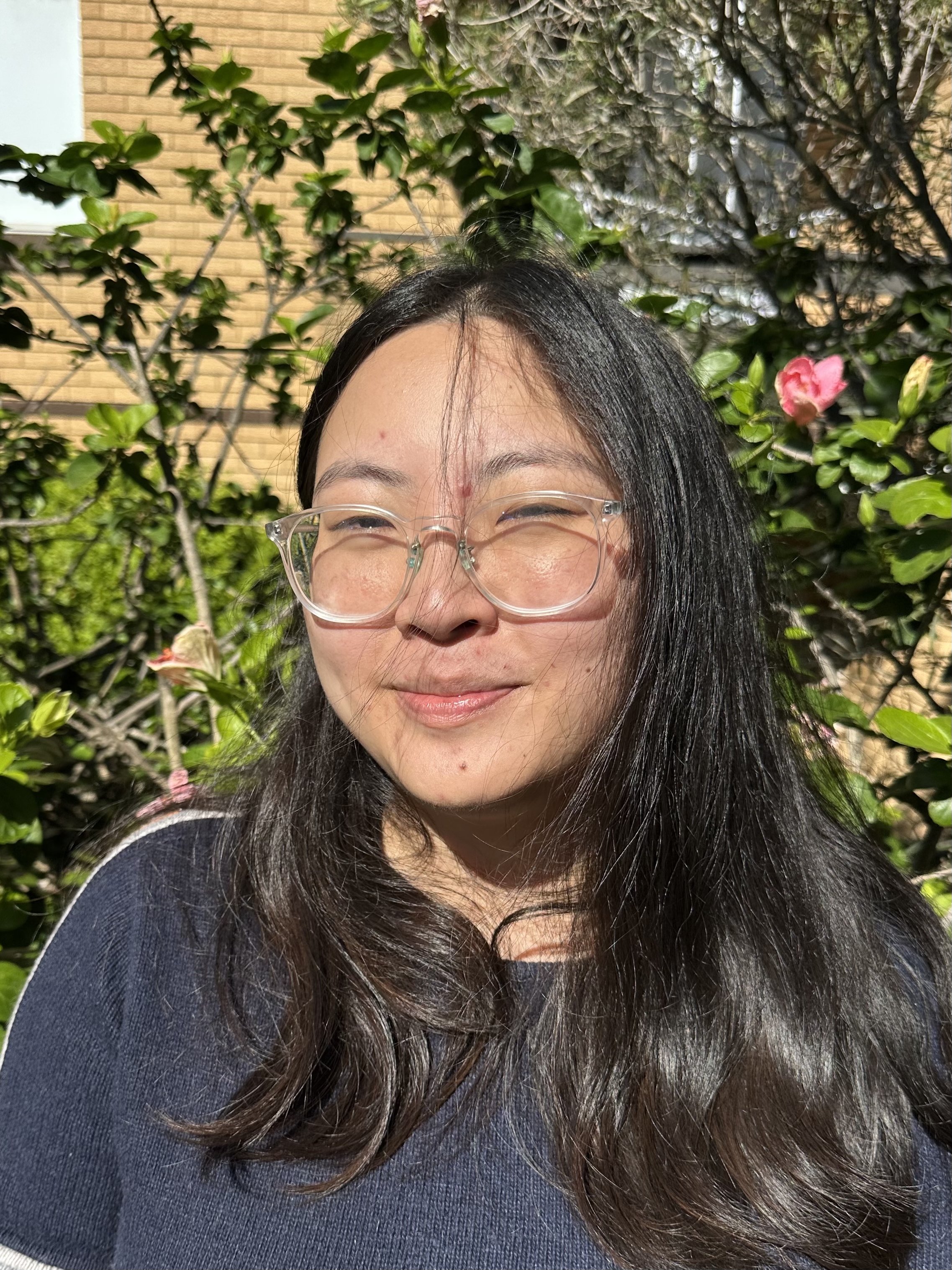 A woman with long black hair, wearing glasses, smiling with her eyes closed, standing outdoors in front of greenery and a brick wall.