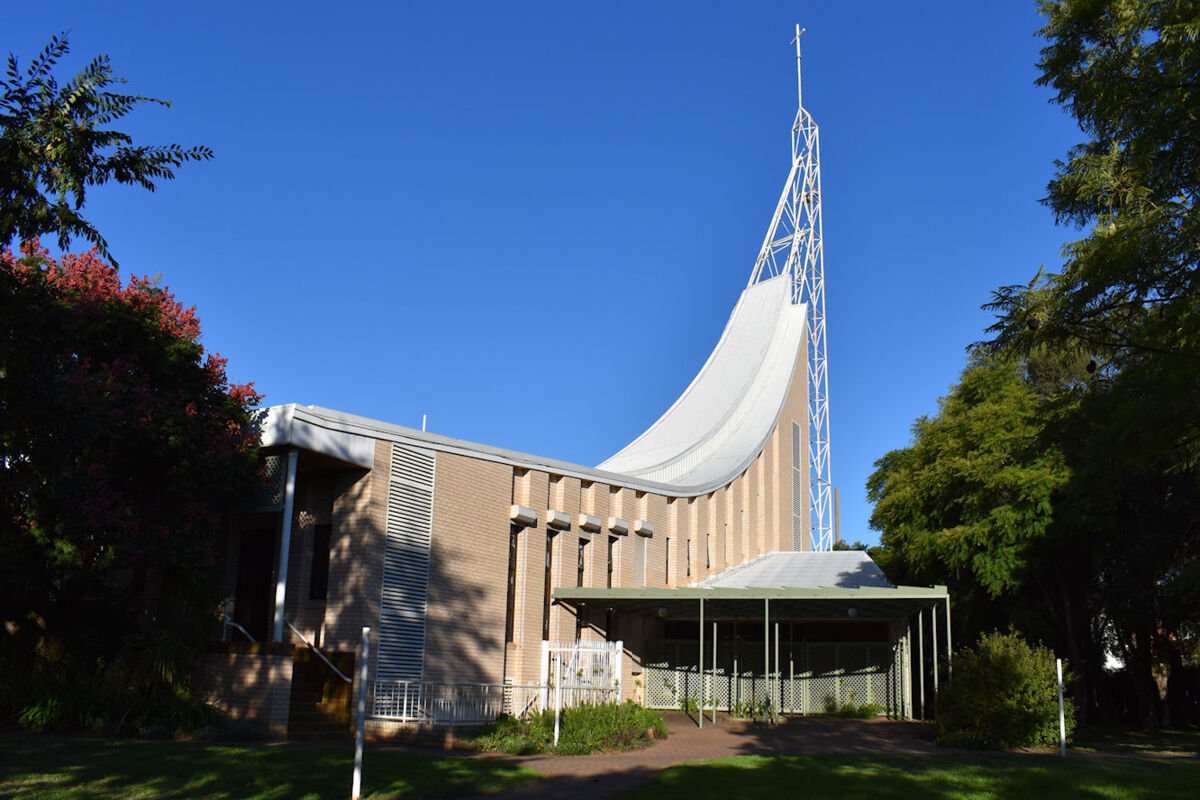 Modern church with a unique curved roof and metal spire against a clear blue sky, surrounded by trees.
