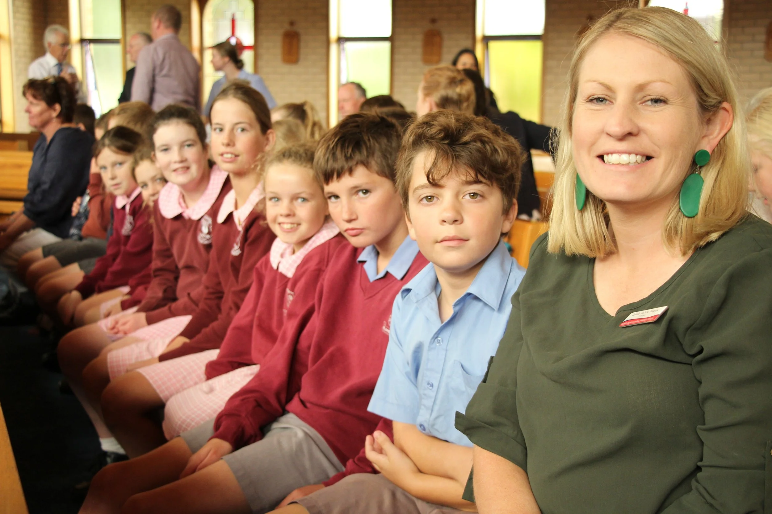 A group of schoolchildren in uniform and a woman sitting in a church setting, smiling at the camera.