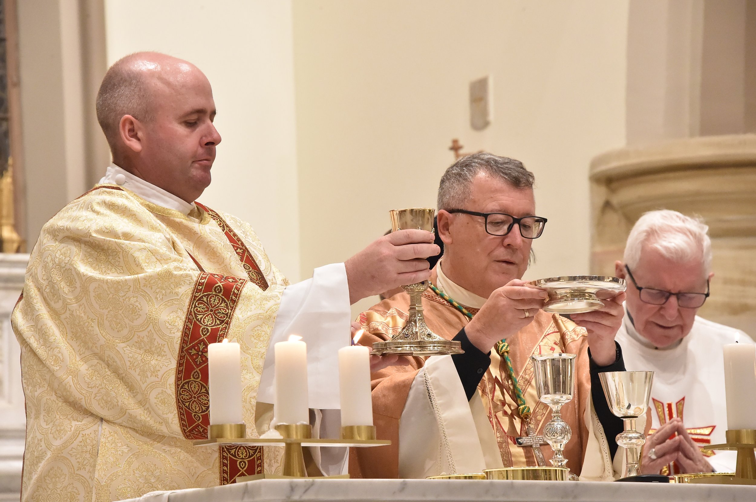 Three priests in ornate vestments holding chalices and patens during a church ceremony, with lit candles on the altar.