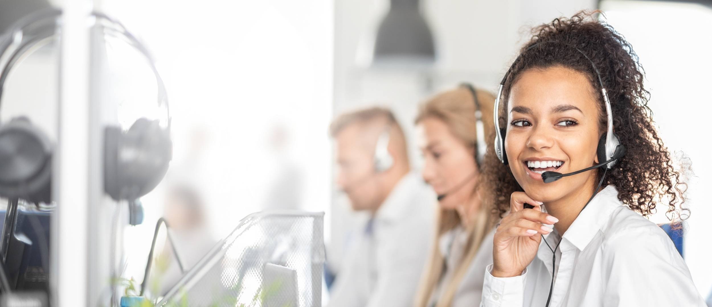 A smiling woman wearing a headset in a call center, with colleagues in the background.