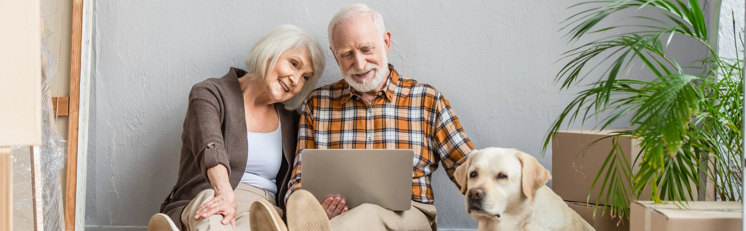 Older couple sitting on floor with laptop, boxes, and dog nearby.