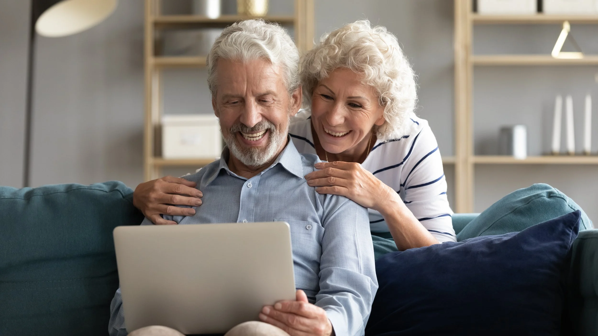 Elderly couple smiling at a laptop screen while sitting on a couch at home.