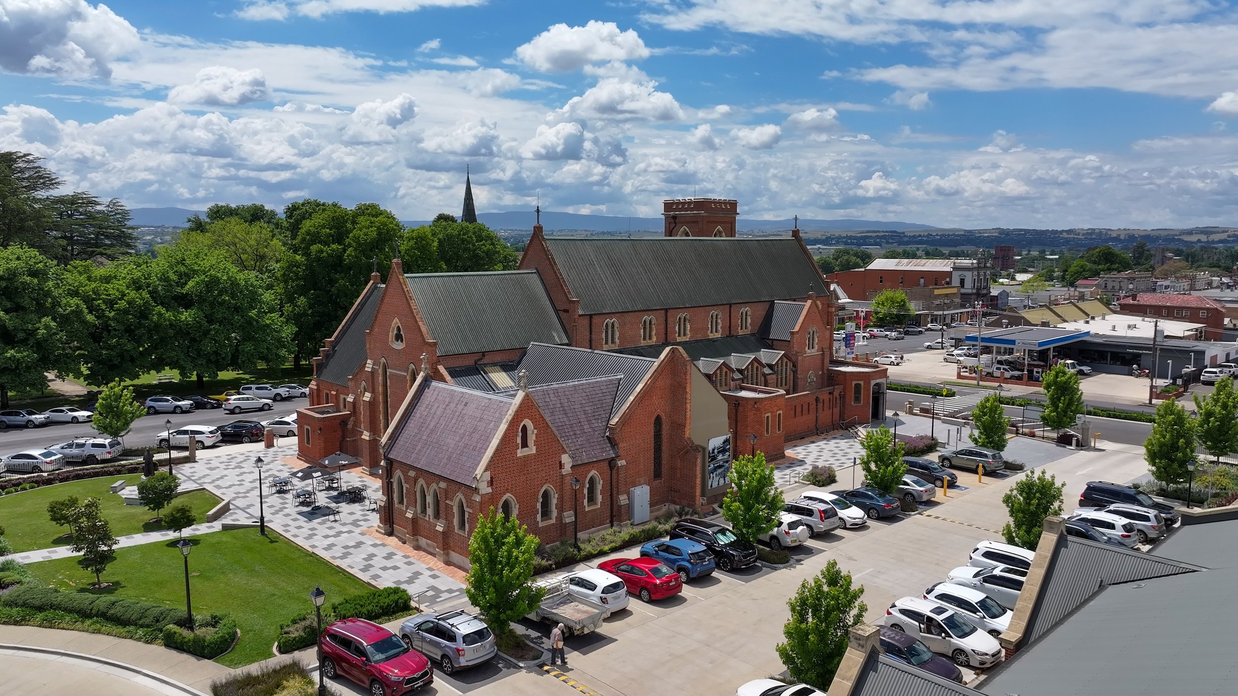 Aerial view of a large brick church building with green roofs and arched windows, surrounded by a parking lot with cars and green trees. The sky is partly cloudy with blue patches.