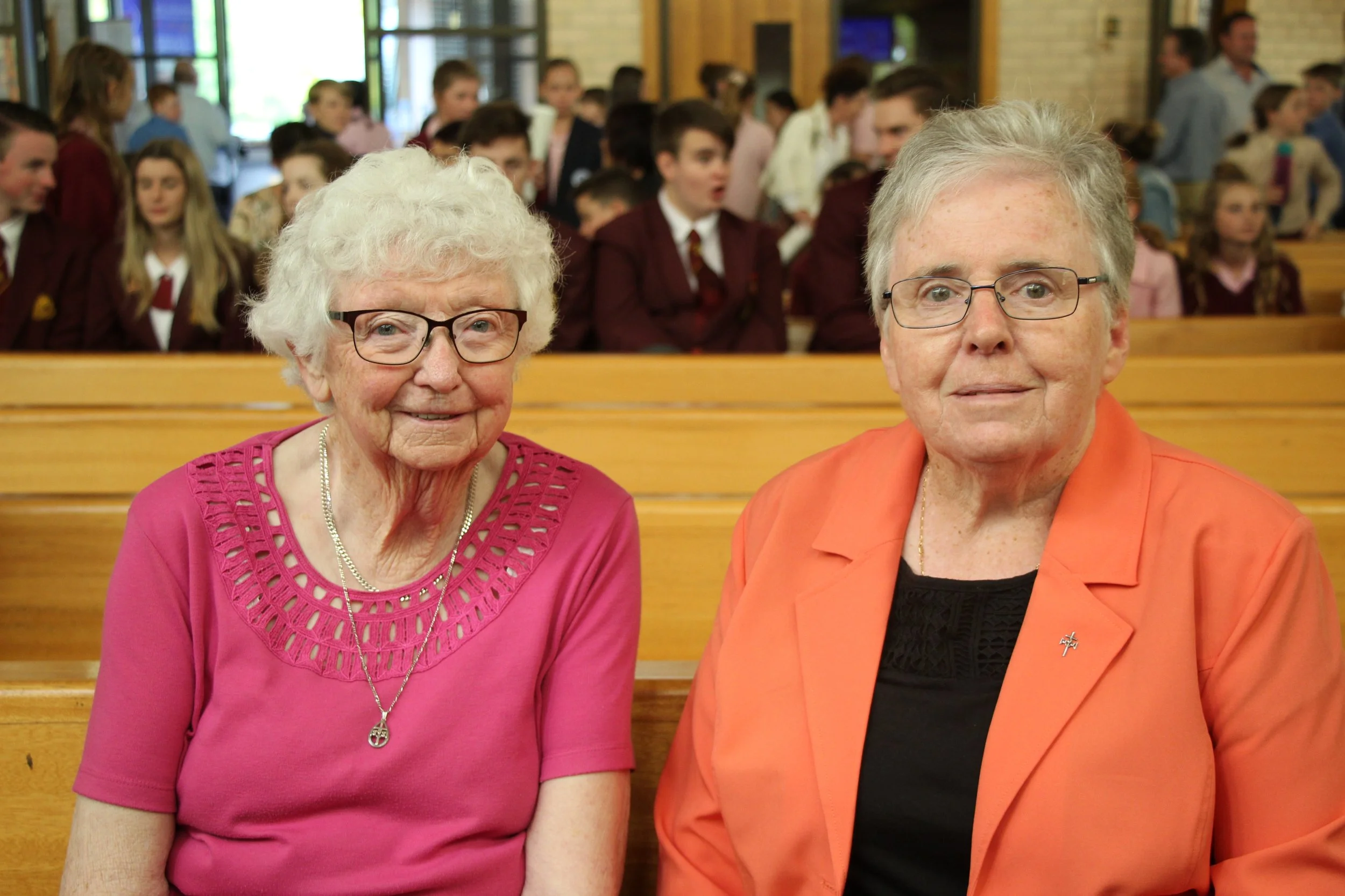 Two elderly women sitting on wooden benches, one wearing a pink top and the other an orange jacket, with people in the background.