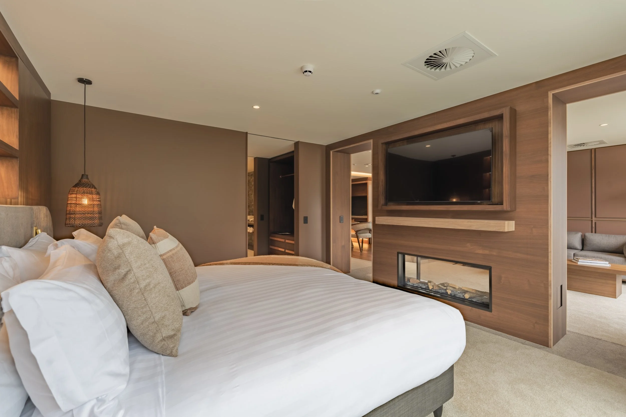 Modern bedroom with a double bed, white bedding, brown blanket, wood paneling, pendant lights, built-in shelves, and an adjoining bathroom.