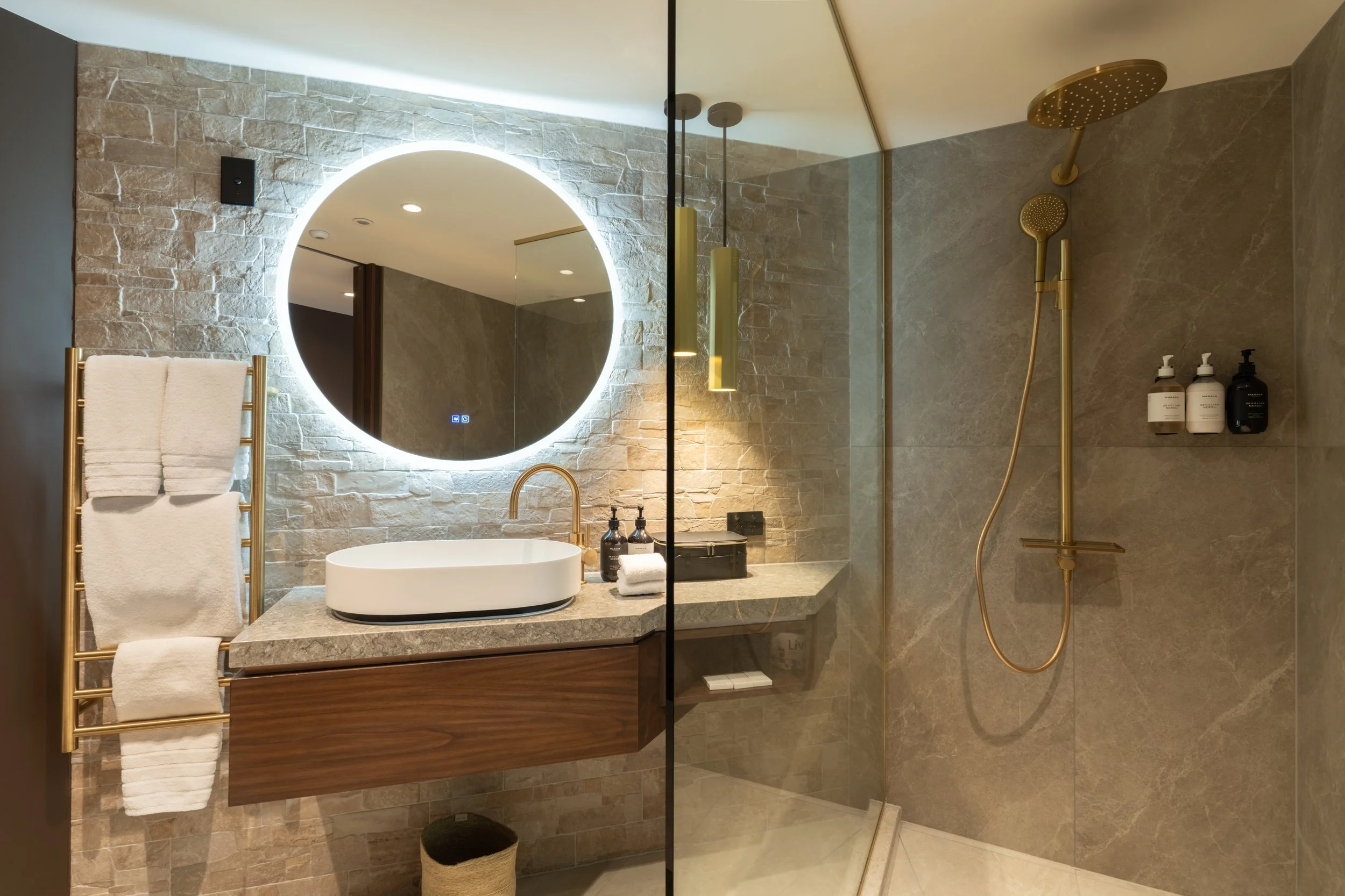 Modern bathroom with stone vanity, round illuminated mirror and walk-in shower.