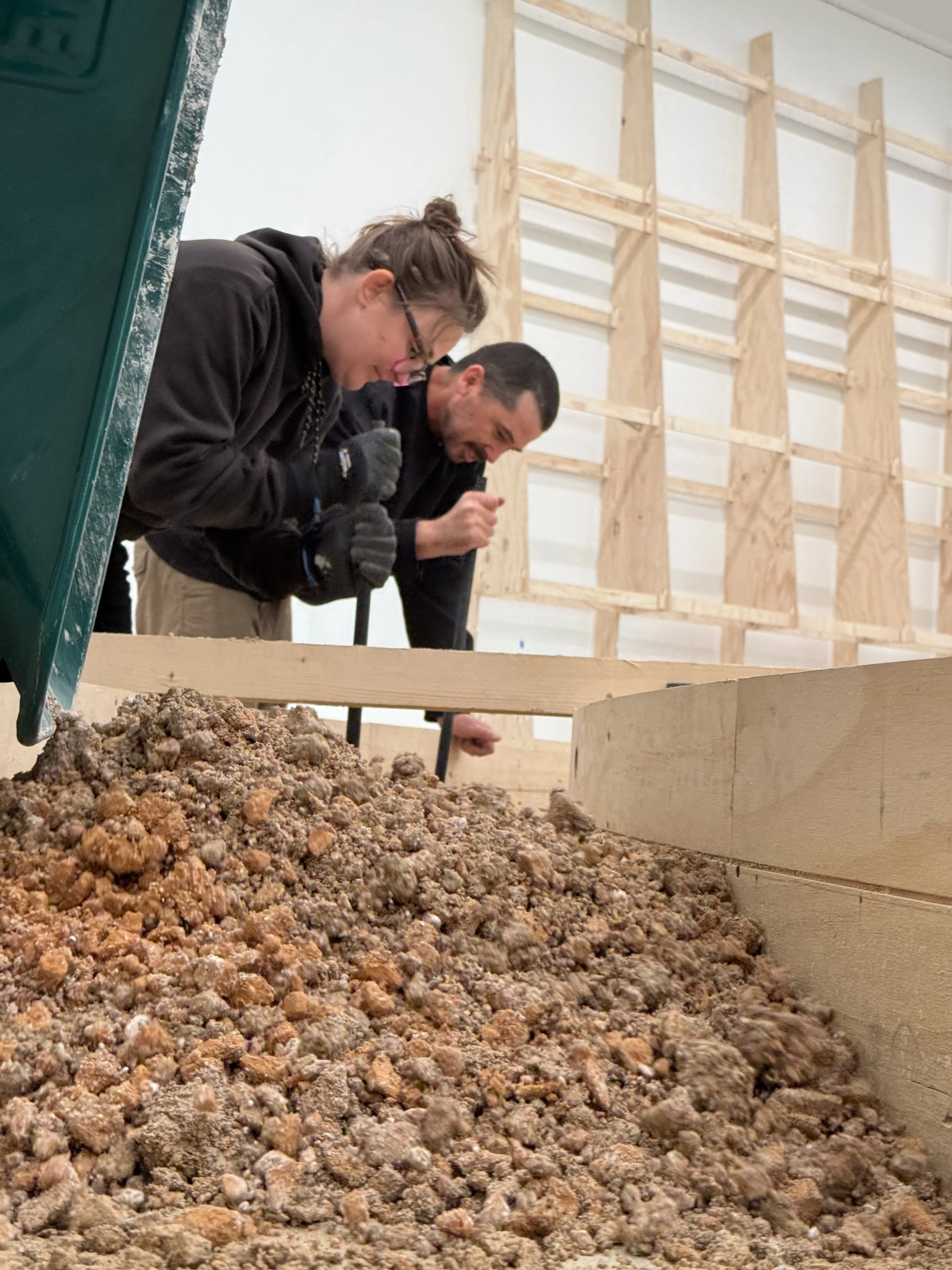 Two people working in a construction or renovation setting, shoveling or moving a pile of dirt or debris into a wooden frame structure. They are indoors, wearing casual work clothing, and there are wooden wall supports in the background.