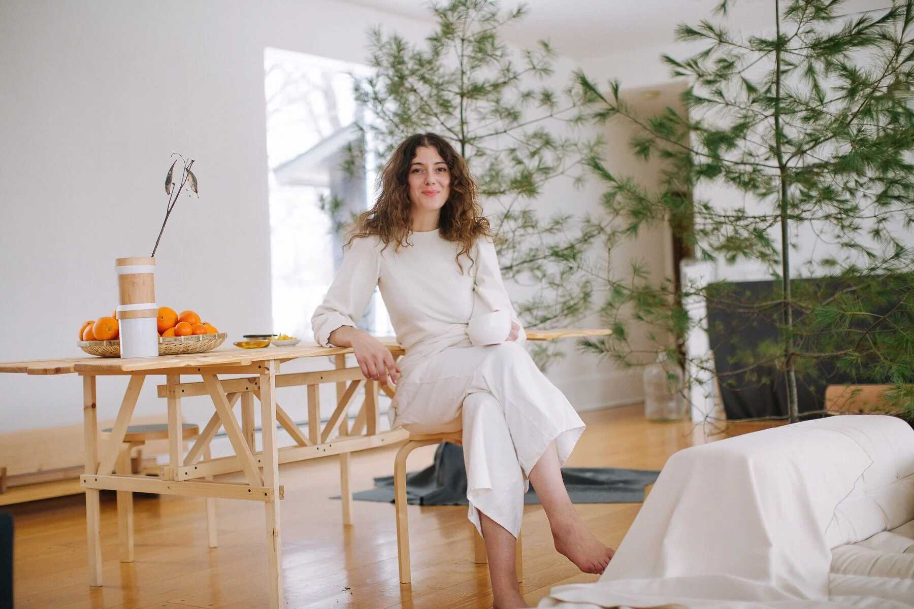 Woman sitting on a chair in a bright room during daytime, holding a white mug, surrounded by potted plants, with a table holding oranges, and a white couch in the foreground.