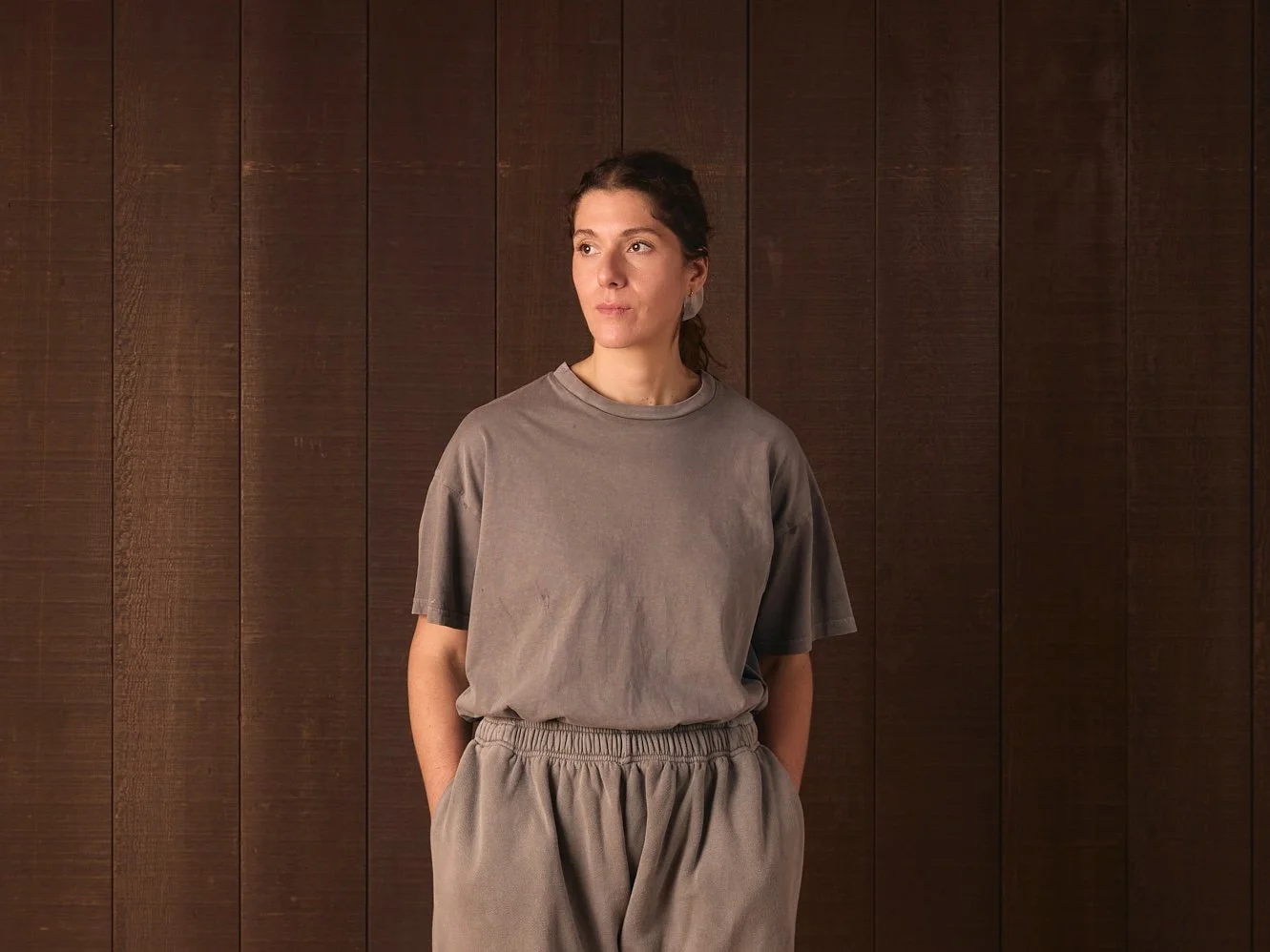A woman standing in front of a brown wooden wall, wearing a relaxed fit gray T-shirt and matching gray sweatpants, with her hands in her pockets.