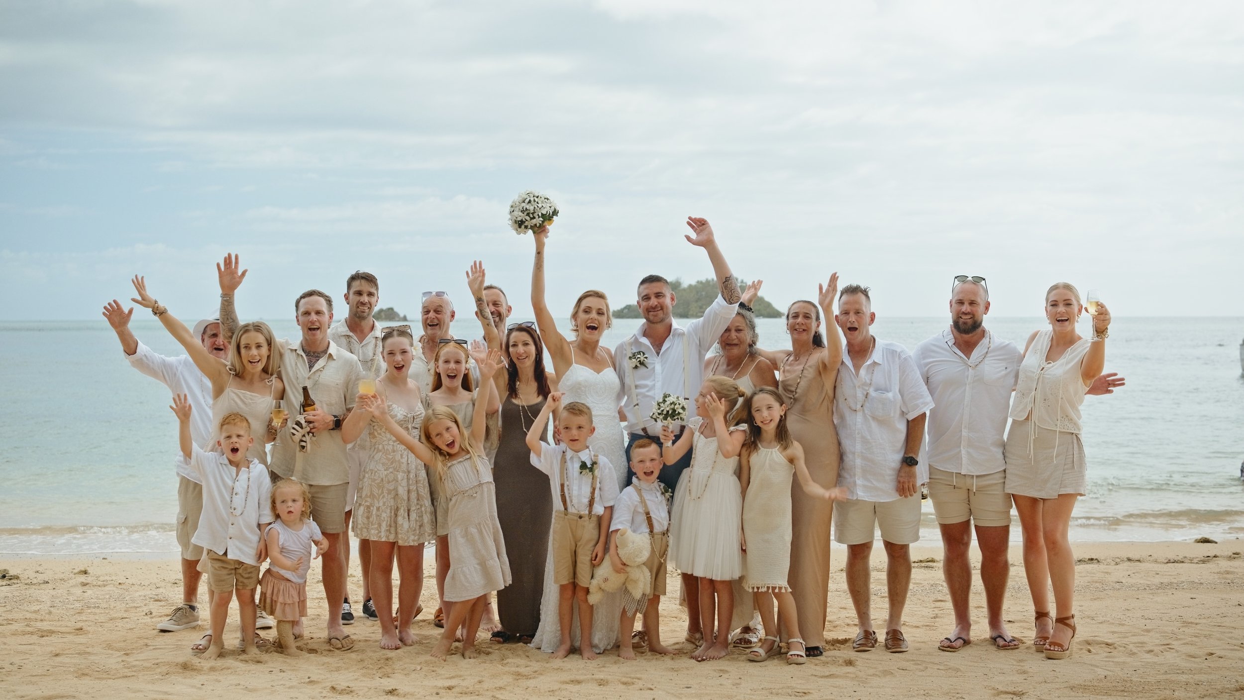 Group of people celebrating a beach wedding, including the bride and groom, children, and family members, on sandy shoreline with the ocean in the background.