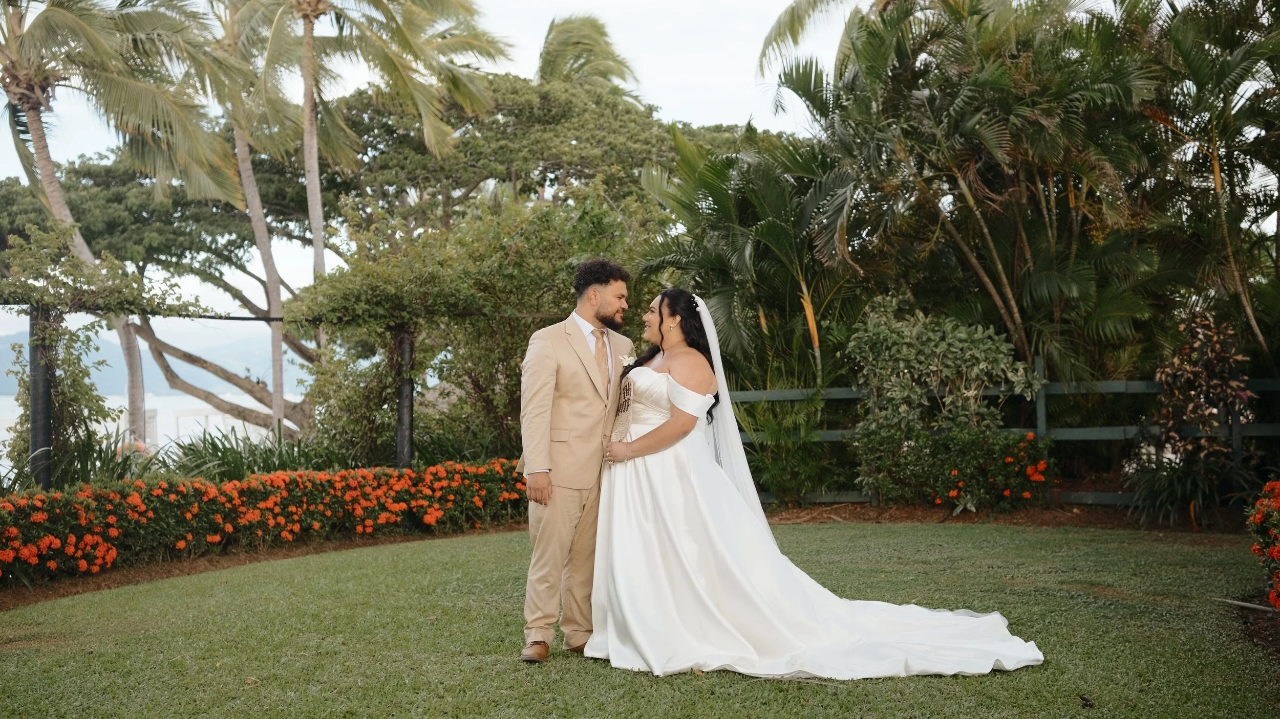 A bride and groom standing and smiling at each other outdoors in a garden surrounded by trees and orange flowers.