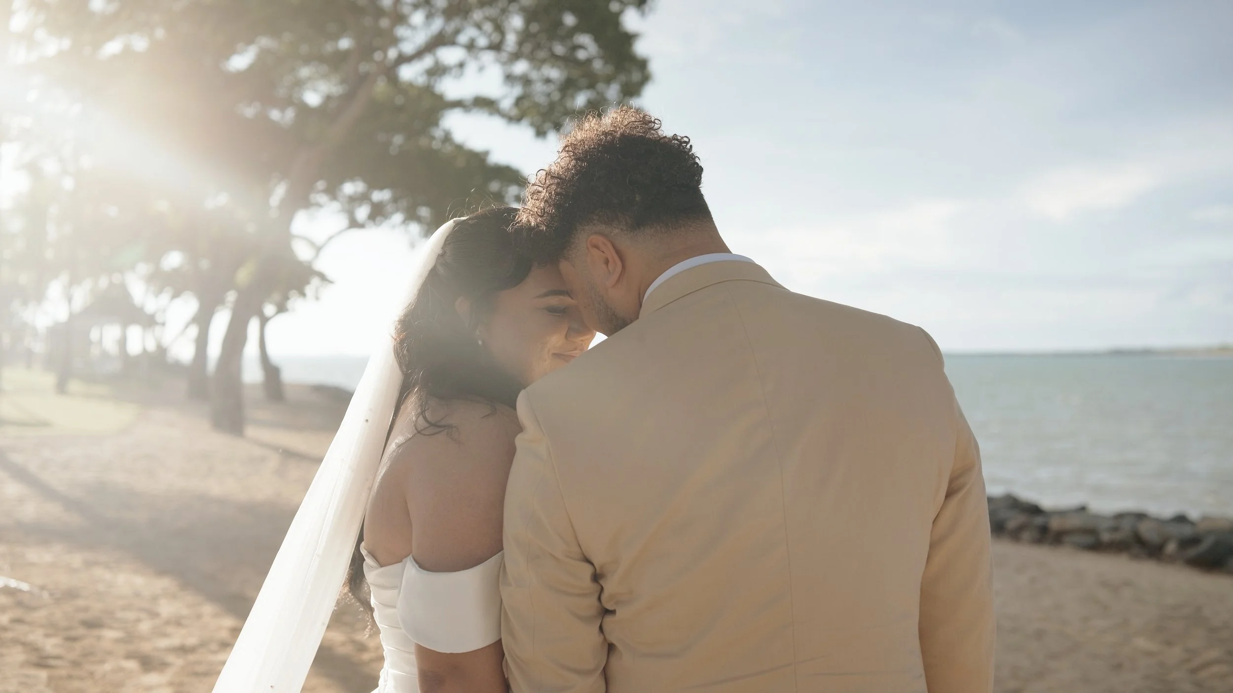 A bride and groom share an intimate moment on a beach, with the bride smiling softly and the couple's foreheads touching, the background features trees and water under a partly cloudy sky.