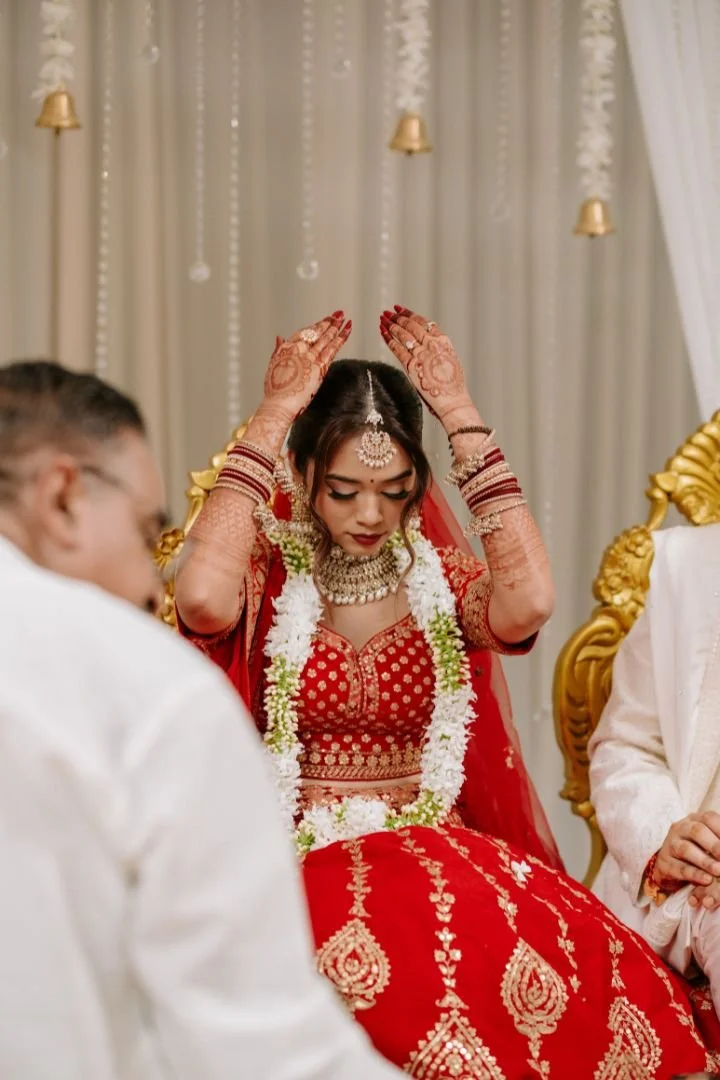 A woman in a traditional red Indian bridal dress is sitting with hands raised, participating in a wedding ceremony, decorated with jewelry and floral garlands, in a decorated indoor setting.