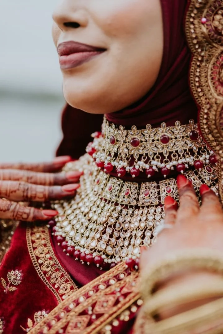 Close-up of a woman wearing traditional red and gold jewelry, including layered necklaces adorned with red and white stones, and a matching embroidered red outfit. She is also wearing henna on her hands.