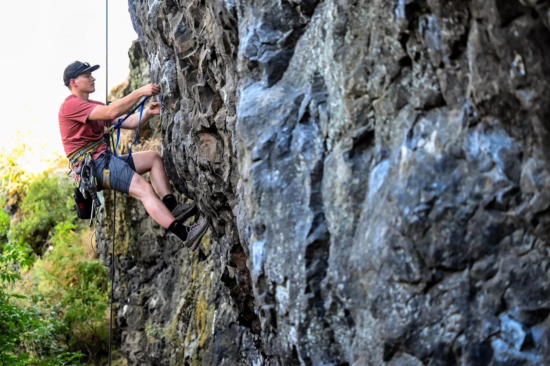 Person replacing climbing bolts on a steep cliff face with safety gear, including a harness and ropes, wearing a red shirt and cap.