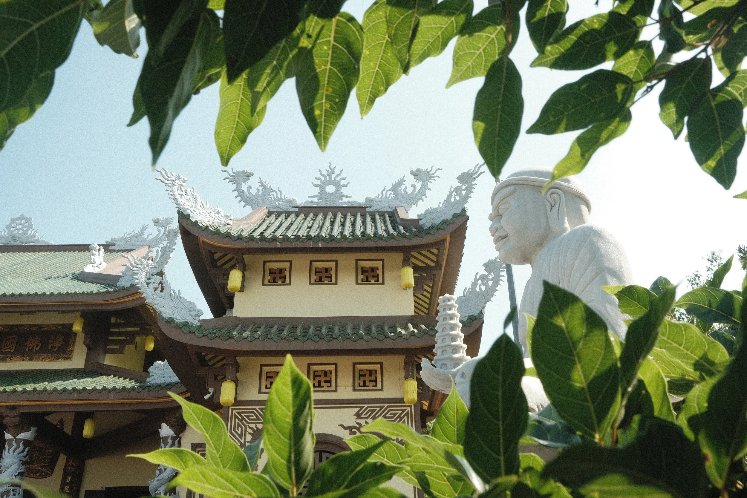 View of a traditional Asian temple with a statue, framed by green leaves.