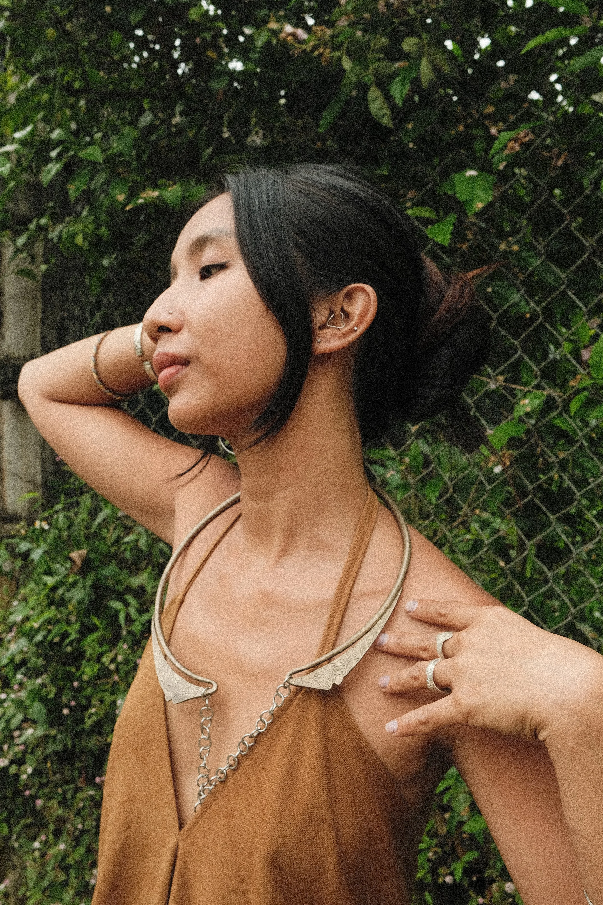 Person posing outdoors, wearing a brown top and silver jewelry, lush greenery in the background.