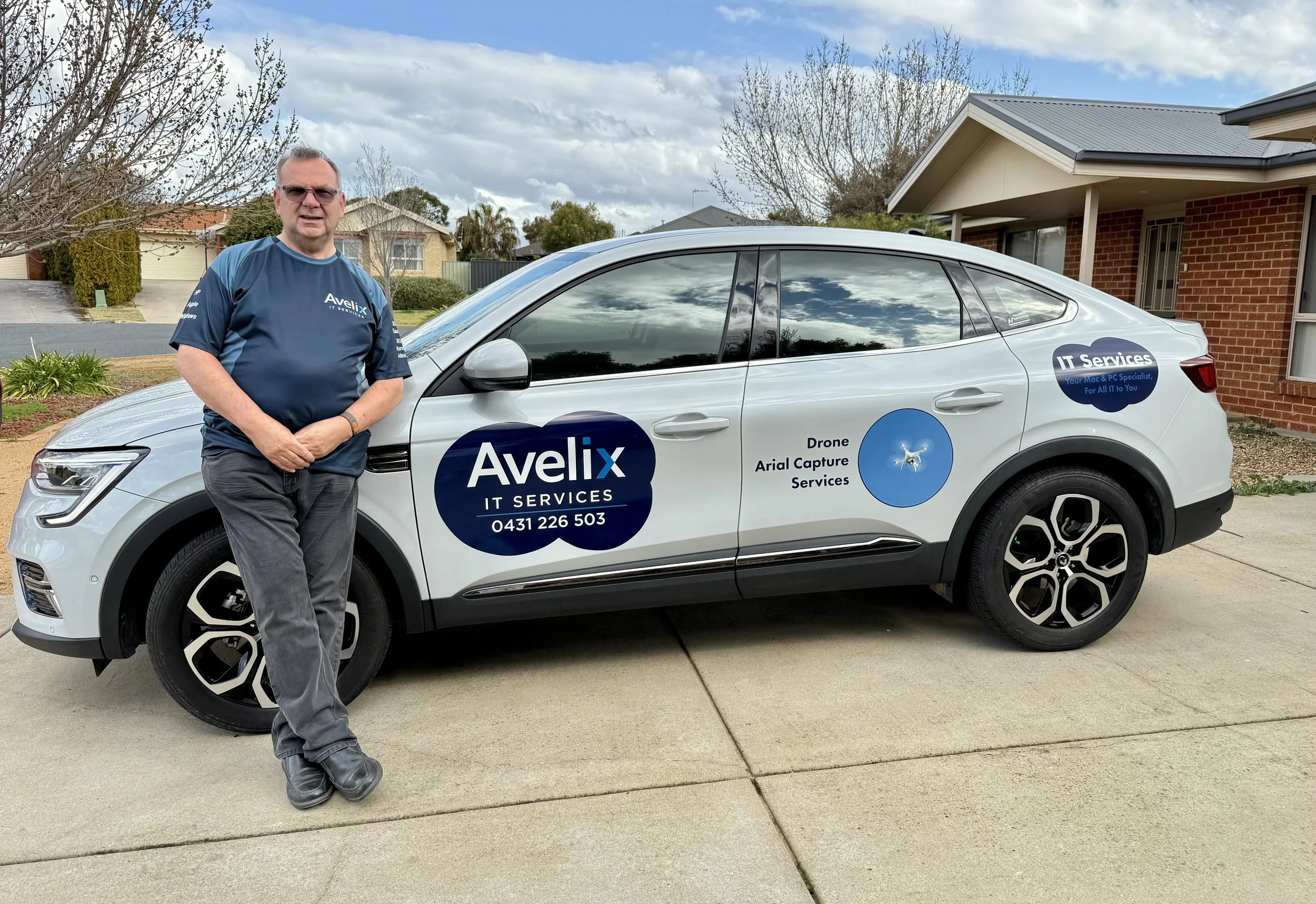 Business owner Tim, standing next to a white car with company logos and text on it, Avelix IT Services, in a residential neighborhood with trees and houses in the background.