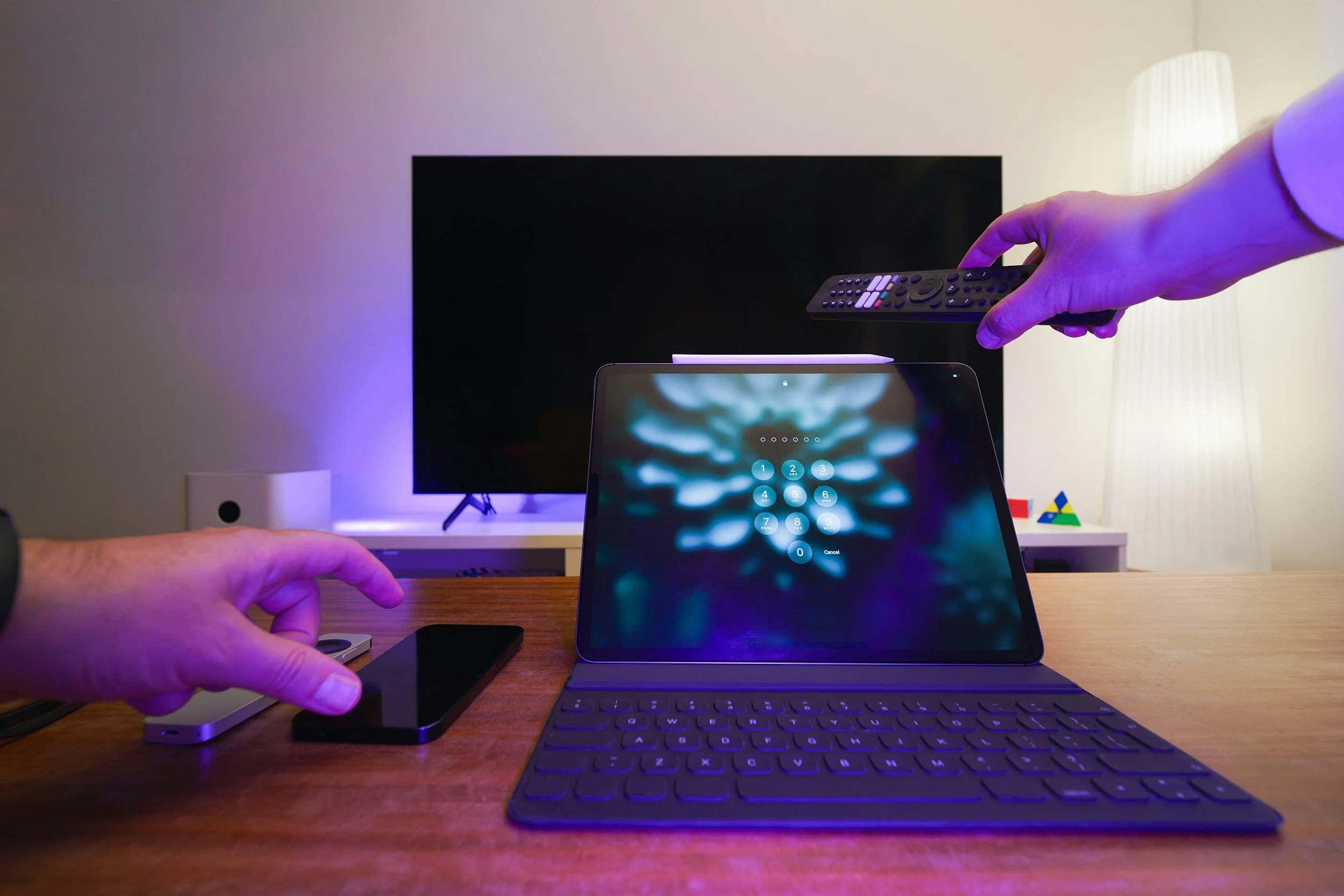 Home office setup with a tablet on a keyboard stand, a TV in the background, a smartphone, and hands interacting with a remote control and the smartphone.