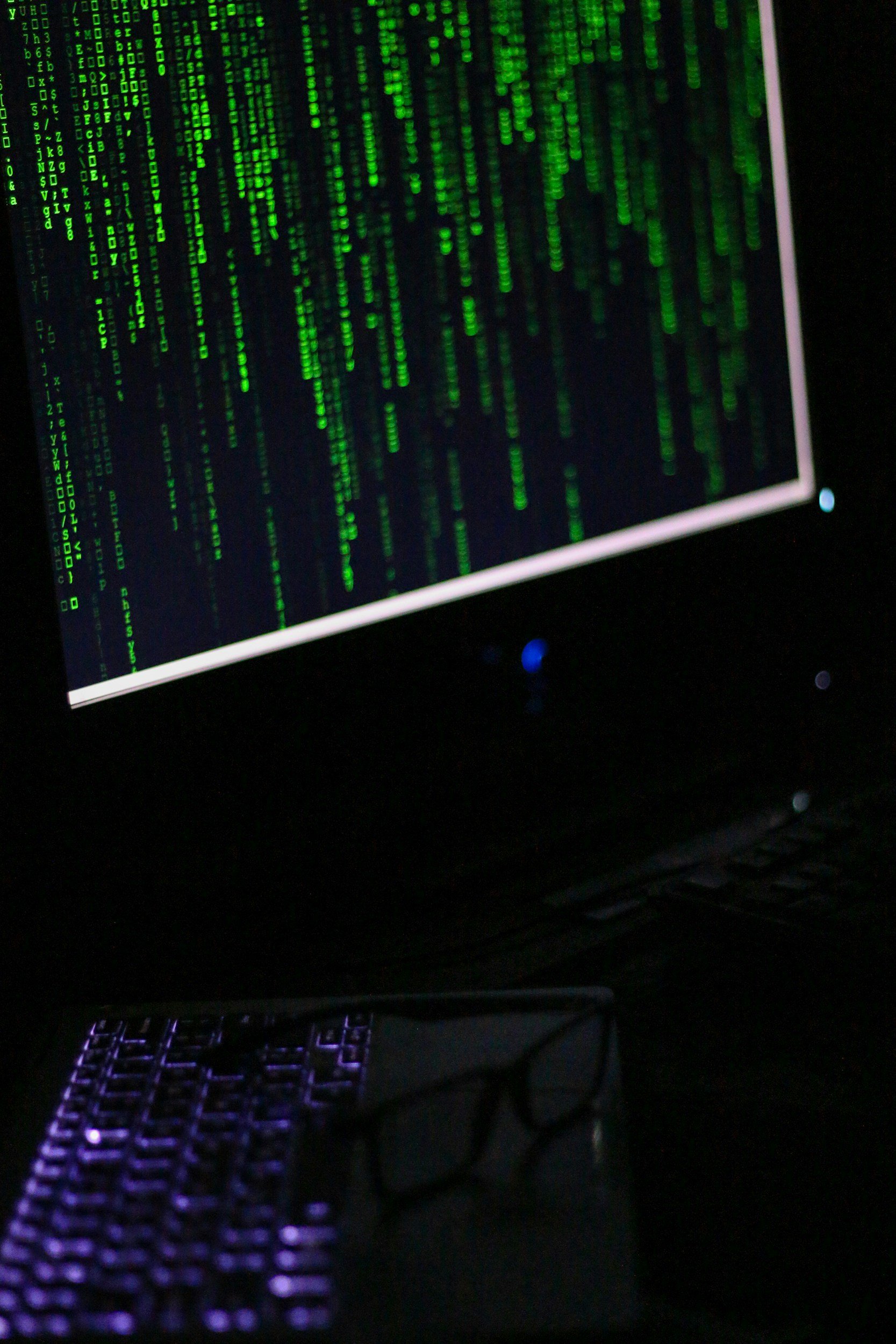 A computer monitor displaying green digital code on a black background, resembling a hacker's screen or coding interface, with a purple-lit keyboard in the foreground.