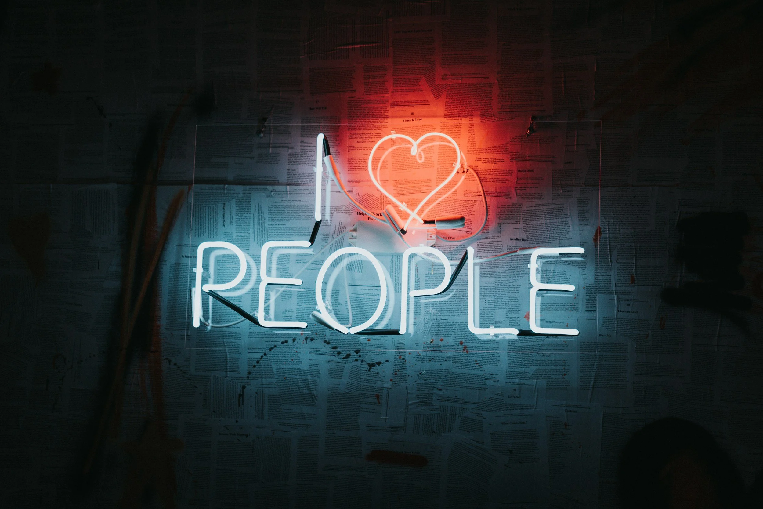Neon sign reading 'I love PEOPLE' with a heart above the word 'love', mounted on a wall covered with newspaper pages.