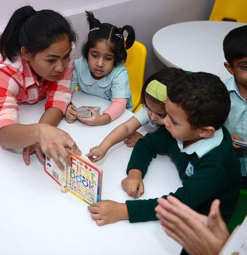 Teacher reading a colorful first word book to young children gathered around a white table in a classroom.