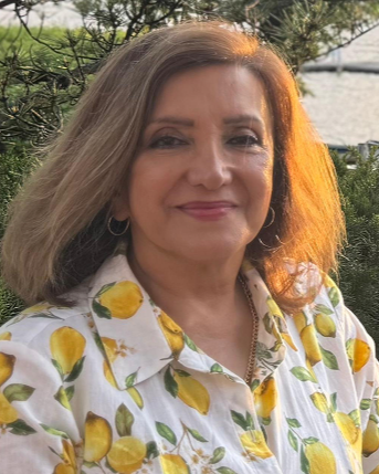 A woman with shoulder-length light brown hair, wearing a white shirt with a lemon pattern, standing outdoors with greenery and trees in the background.