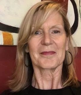 Portrait of a woman with shoulder-length light blonde hair, wearing earrings, in front of a colored background.