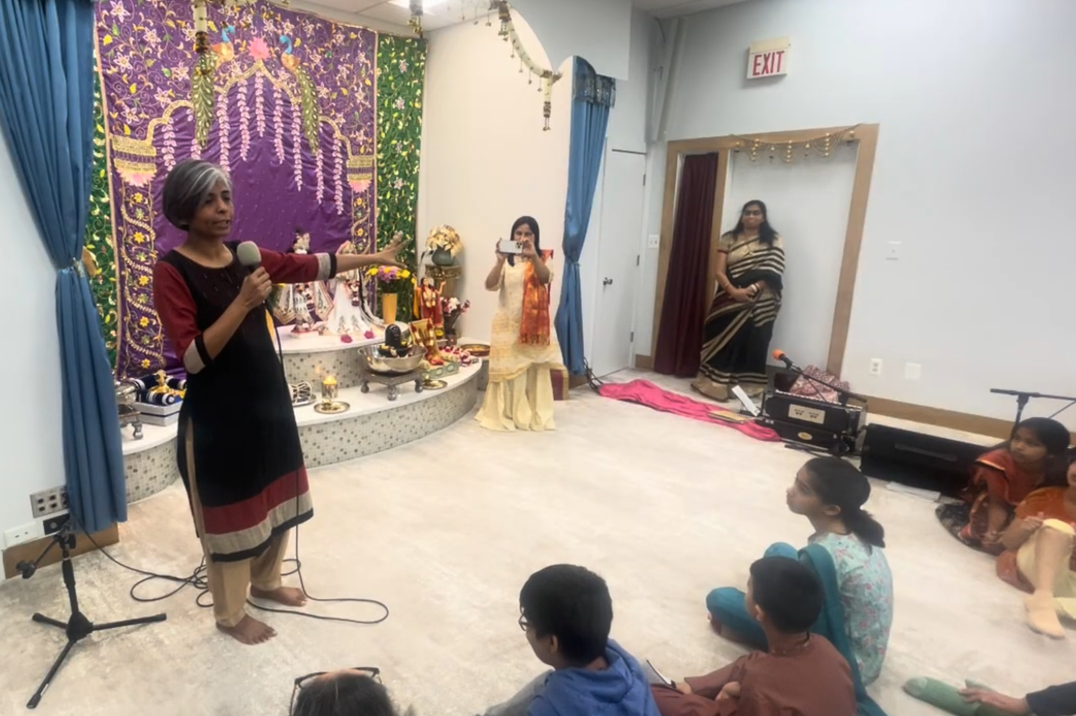 A woman with gray hair and traditional attire is speaking into a microphone while pointing towards a decorated altar with religious and cultural items. Several children are seated on the floor, attentively listening to her. Another woman is taking a photo, and a woman in a striped saree is standing near the doorway, observing the scene. The setting appears to be a cultural or religious event with colorful decorations and an altar in the background.