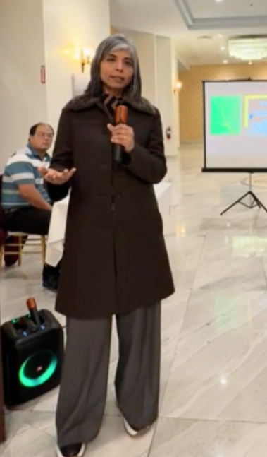 A woman wearing a dark coat and gray pants holding a microphone and speaking at an indoor event with a presentation screen in the background and a man sitting nearby.