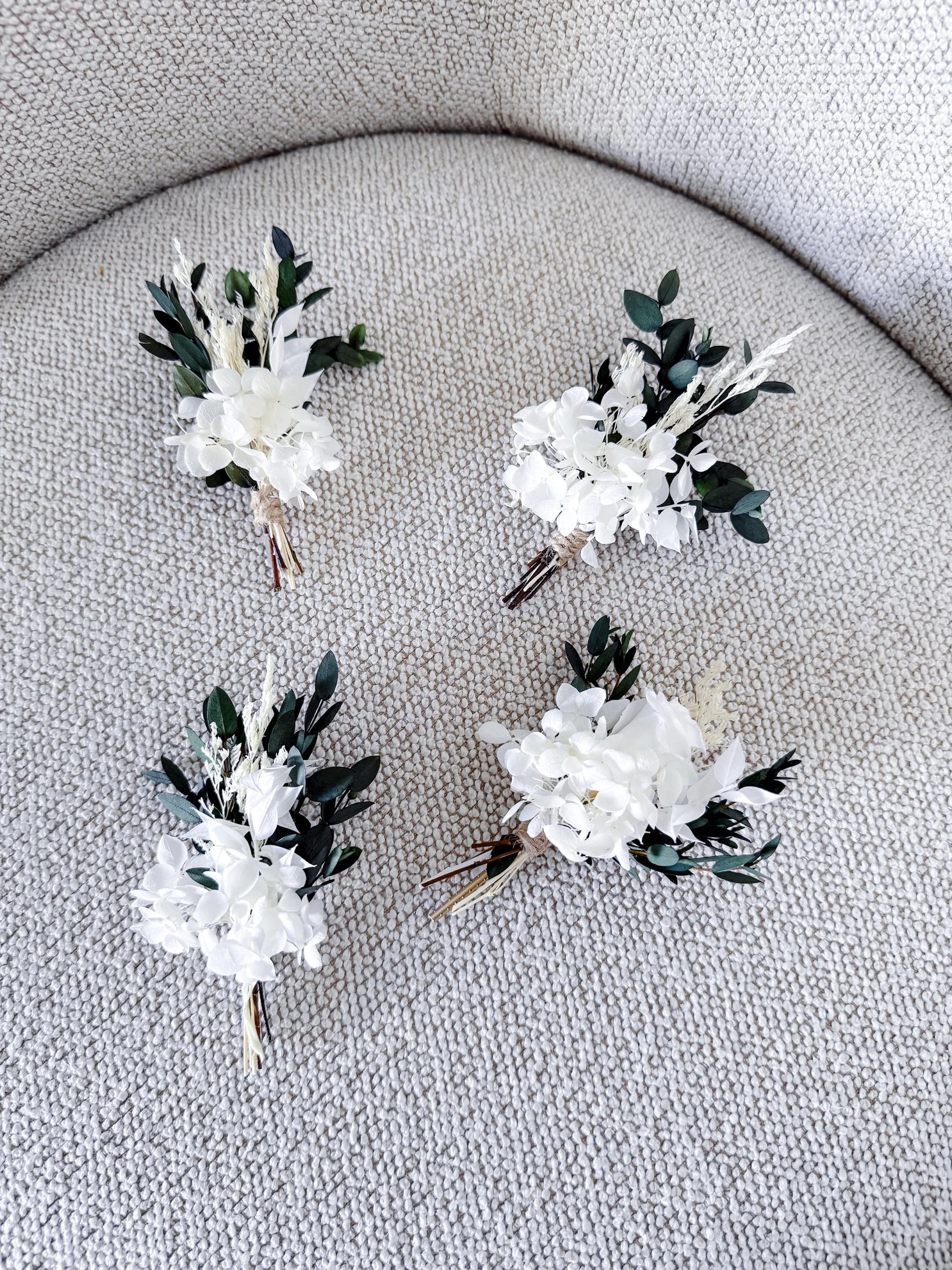 Four white floral boutonnieres with dark green leaves are arranged on a beige textured surface.