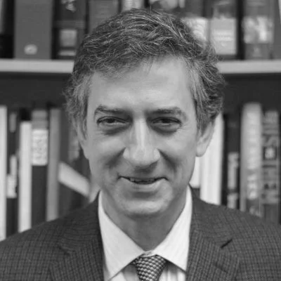 A black and white portrait of a man with tousled hair, wearing a suit and tie, standing in front of a bookshelf.