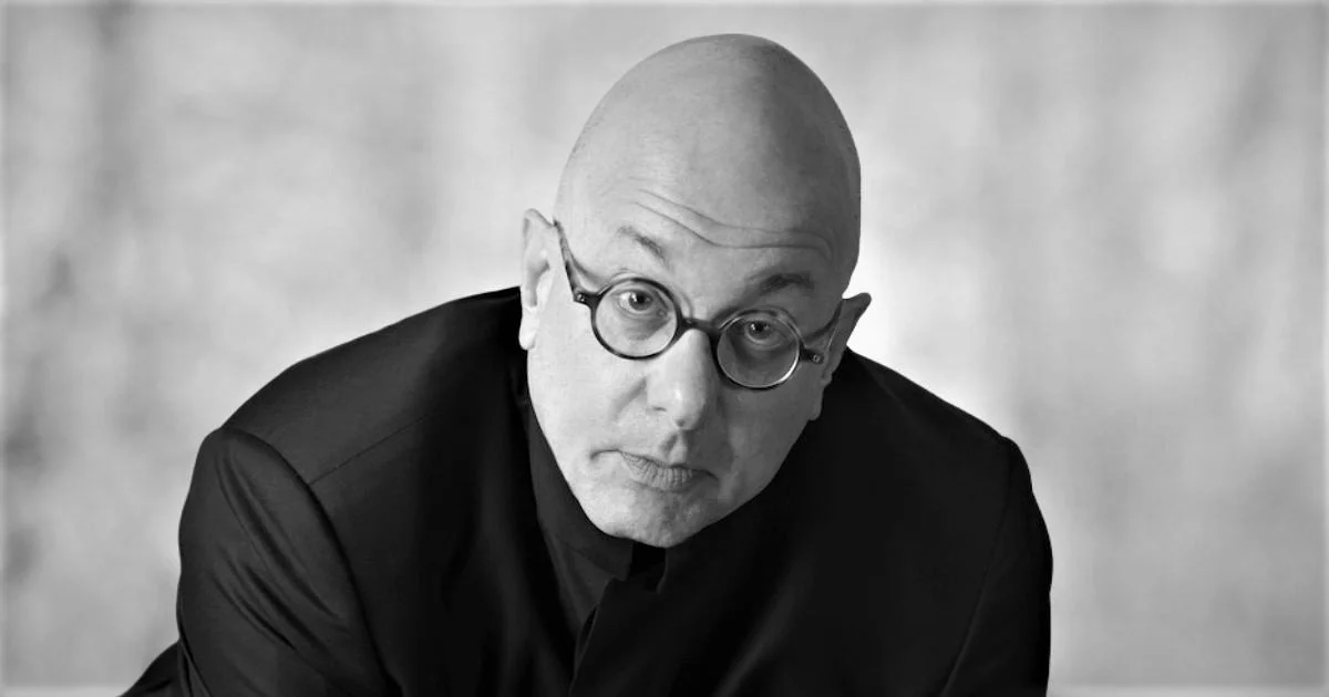 Black and white portrait of a bald man wearing glasses and a dark suit, looking directly at the camera with a serious expression.