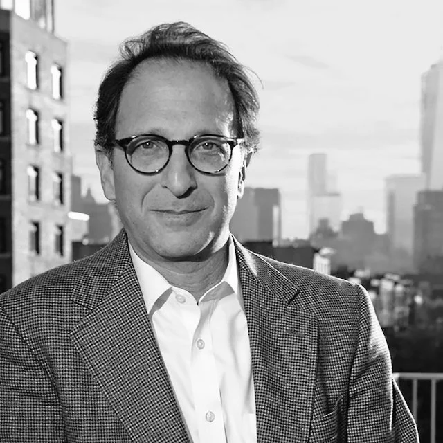 A man with glasses in a suit jacket and collared shirt standing outdoors with a city skyline in the background.