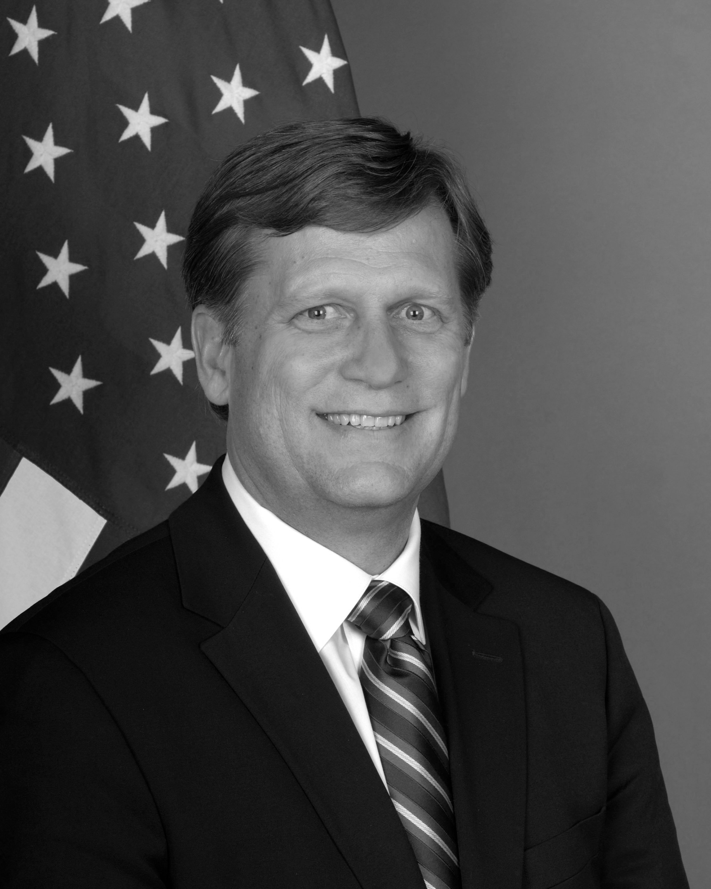 Black and white photograph of a man in a suit and tie, smiling, with an American flag in the background.