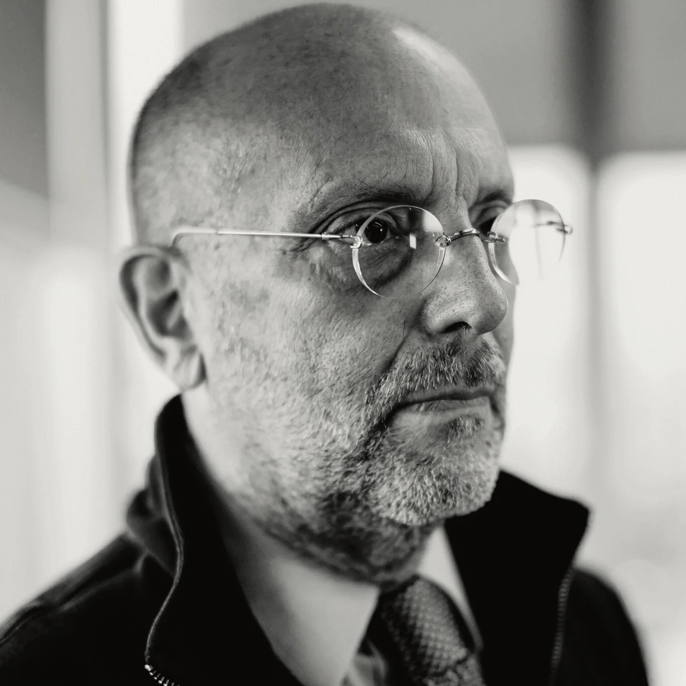A black and white close-up portrait of a man with glasses, a beard, wearing a jacket and tie, looking to the side.
