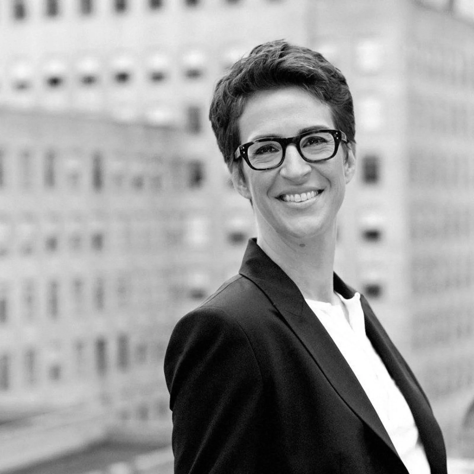 A black-and-white photograph of a smiling woman with short hair and glasses, wearing a blazer, in an urban setting with a building in the background.